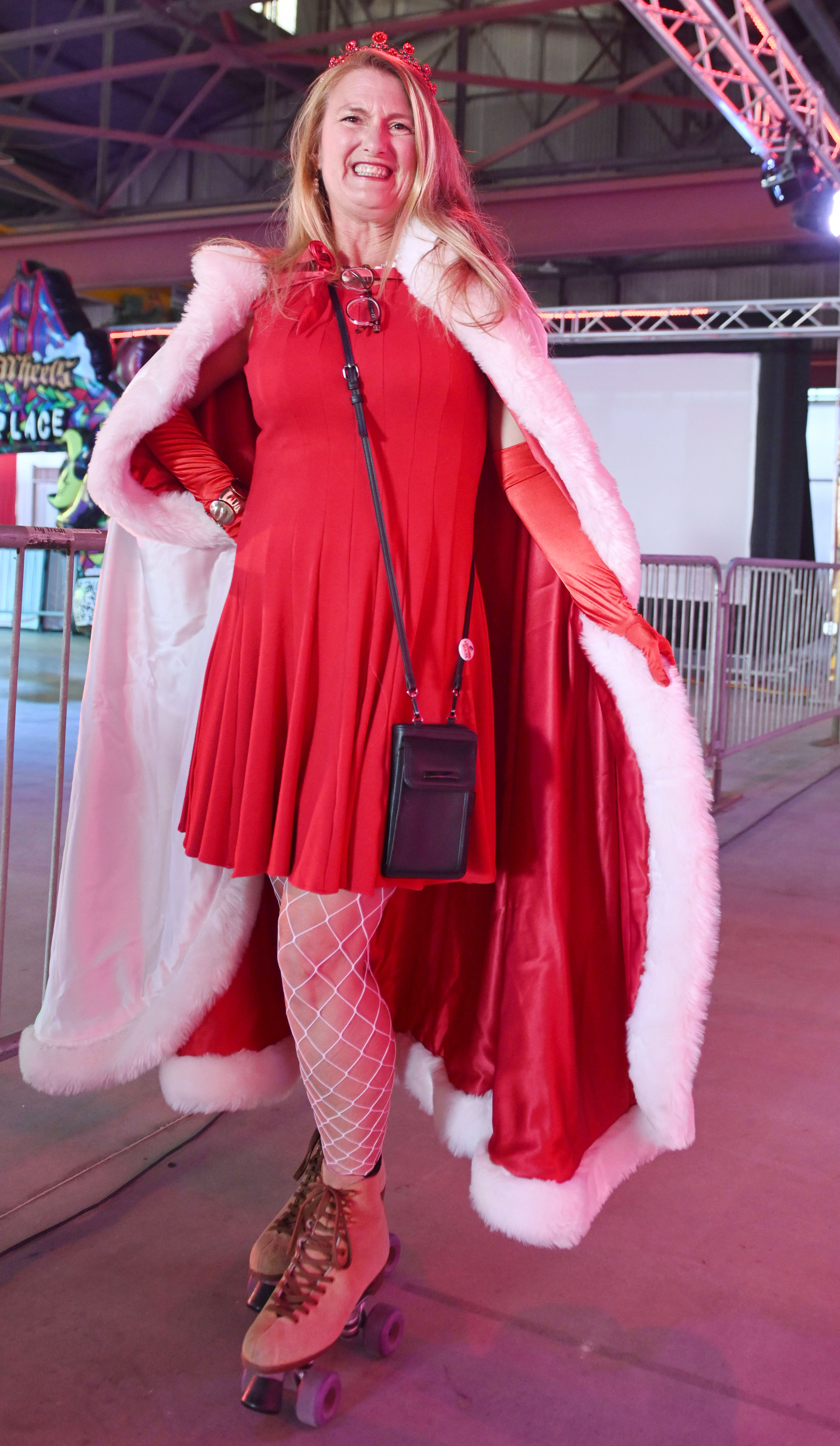 A woman in a red dress and matching cape with white fur trim, wearing fishnet stockings and roller skates, smiles broadly indoors under colored lighting.