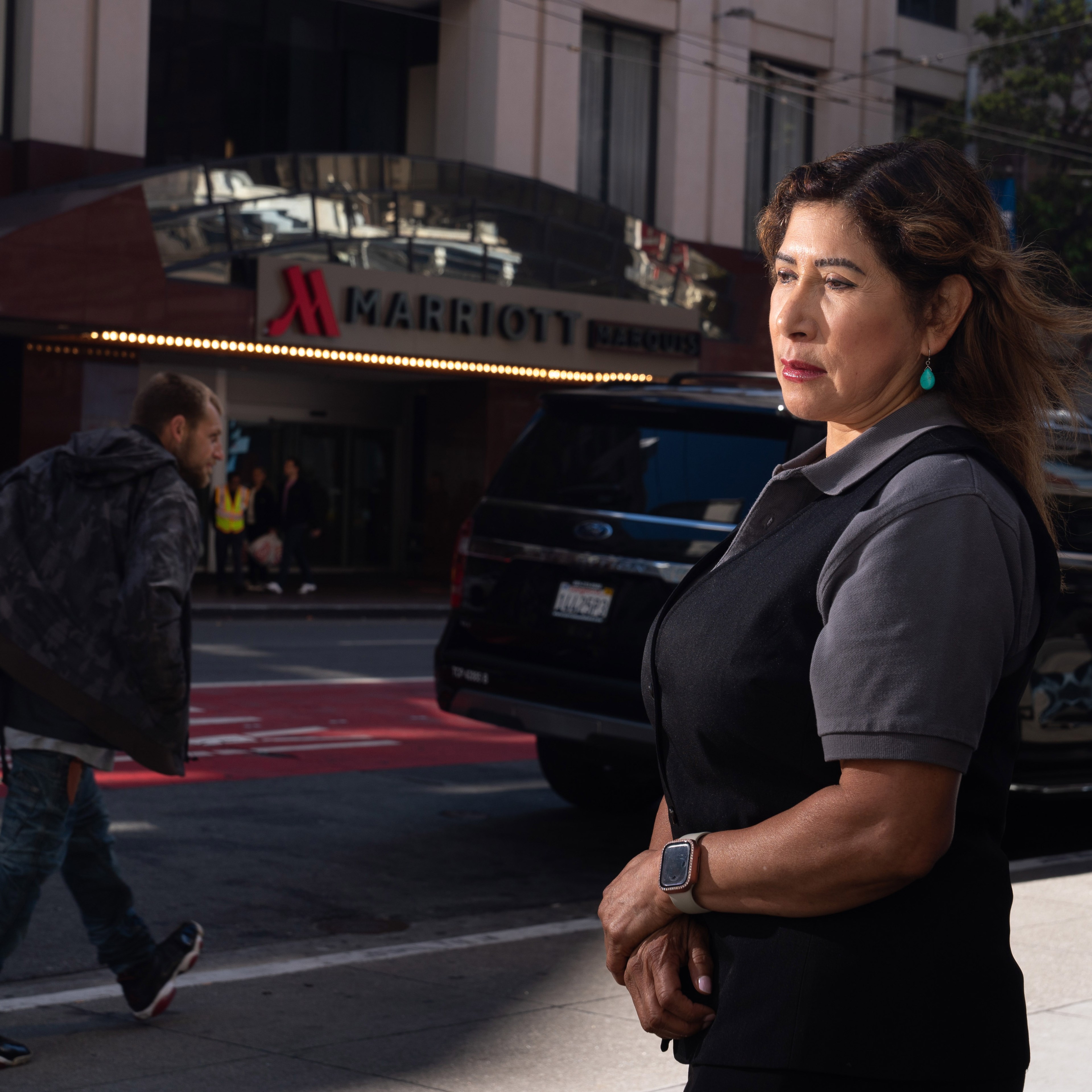 A woman wearing a black vest stands solemnly on a city street, with a Marriott Marquis hotel entrance and a passing pedestrian in the background.