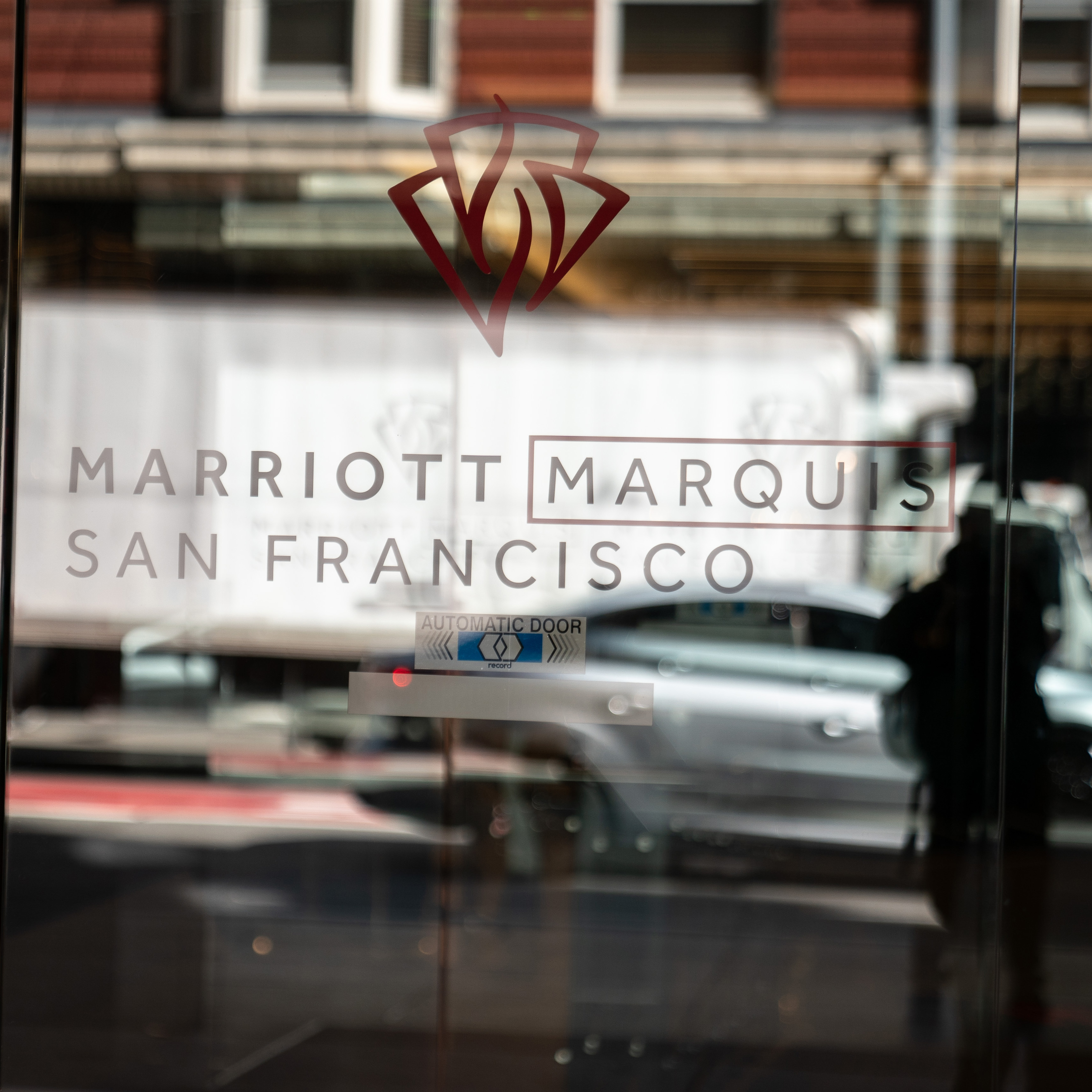 The image shows the glass entrance door of the Marriott Marquis San Francisco hotel, with the hotel name and logo visible, reflecting a street scene outside.