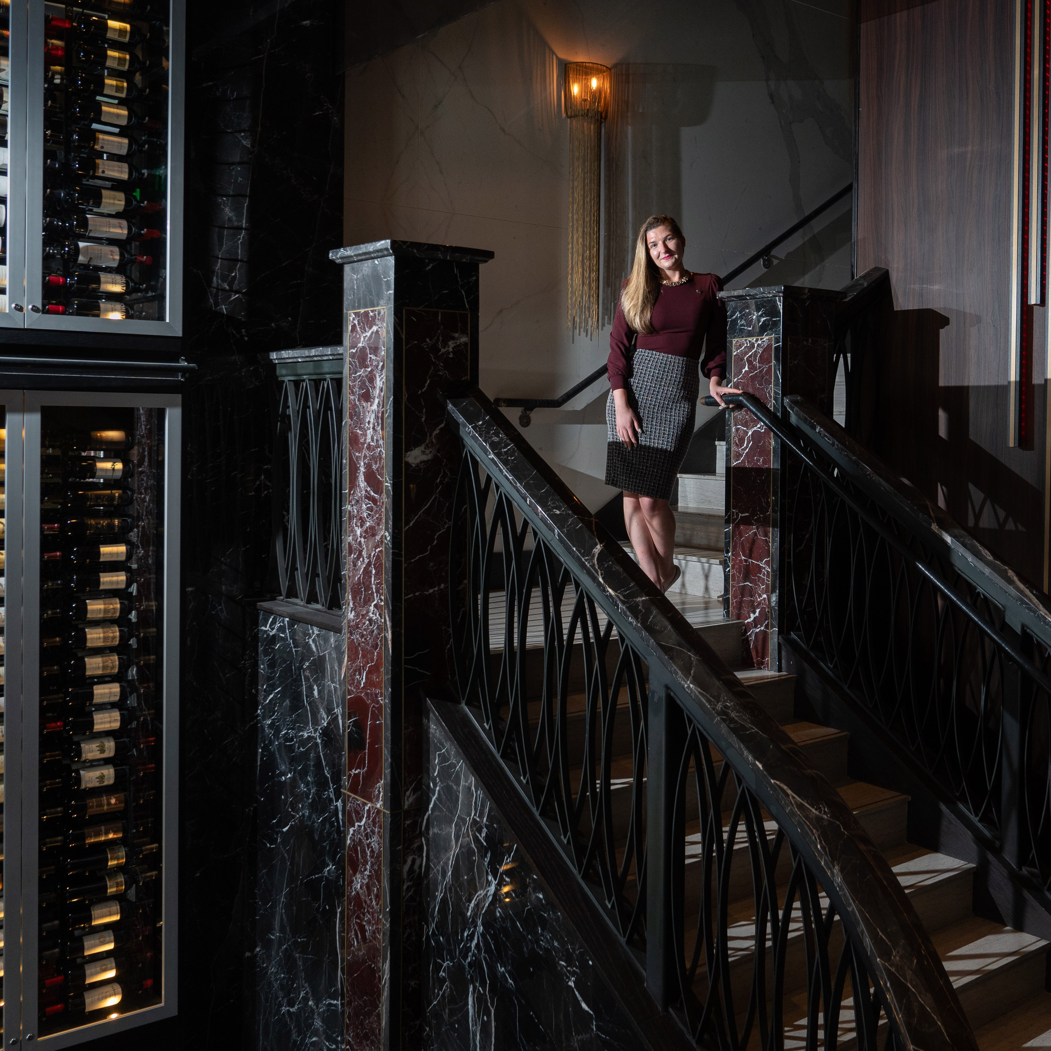 A woman stands halfway down a grand staircase with black marble details, next to a large wine storage display filled with bottles, under a warm light fixture.