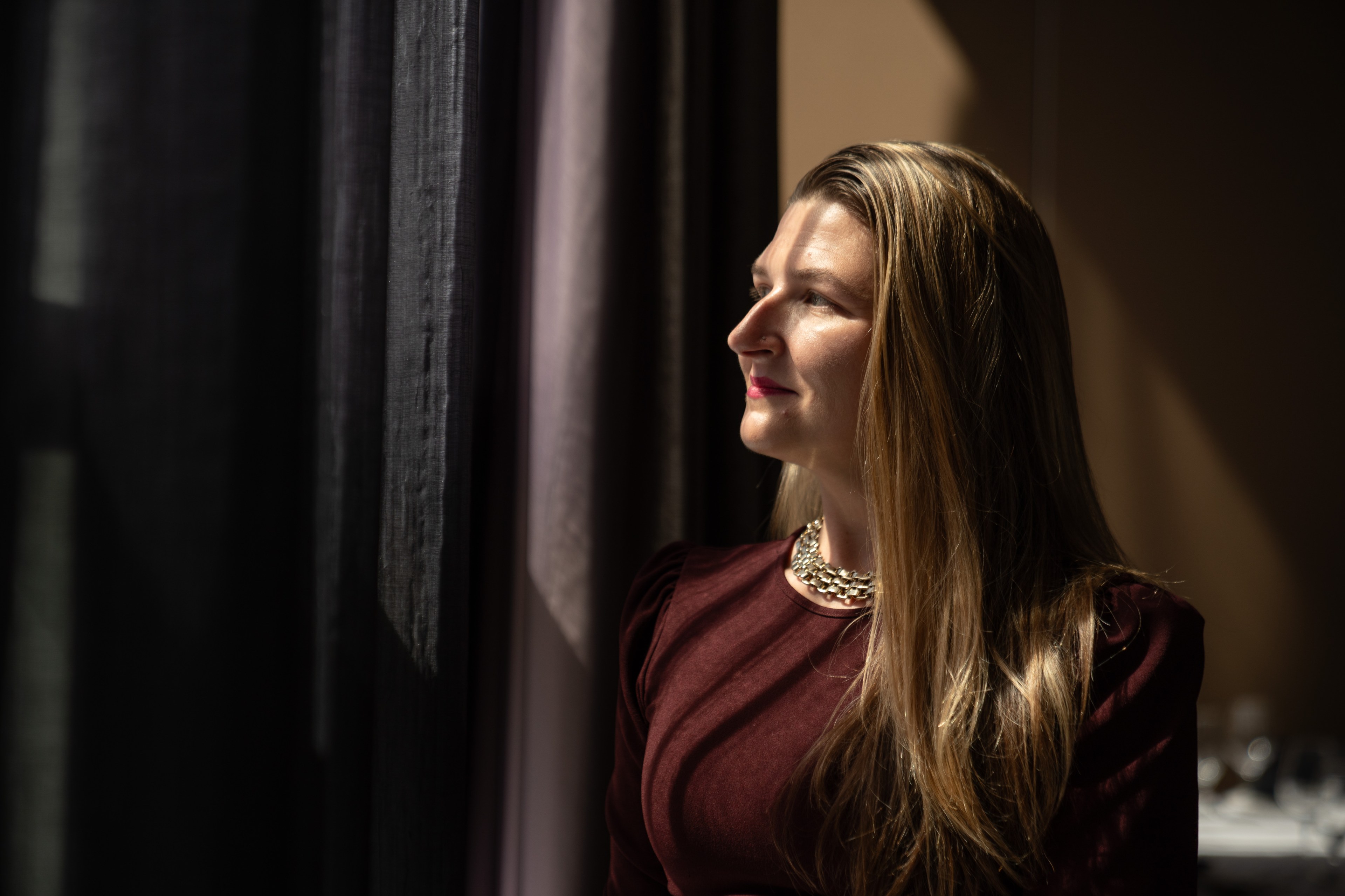 A woman with long blonde hair, wearing a burgundy top and gold necklace, is gazing towards a window with sunlight illuminating her face against a dark curtain backdrop.