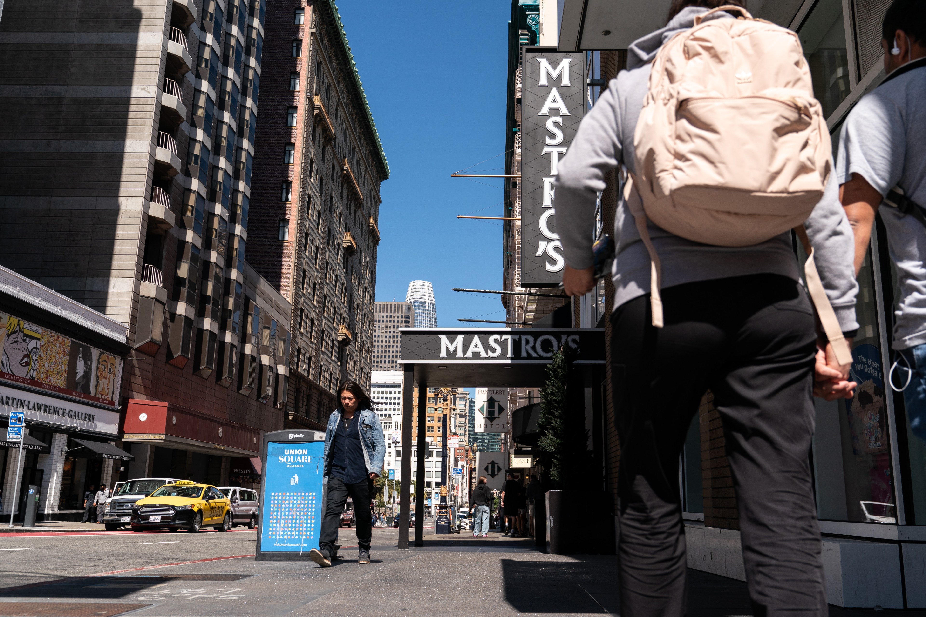 A city street features tall buildings and a blue sky. People walk by, including one person wearing a large beige backpack. A &quot;Mastro's&quot; restaurant sign is visible.