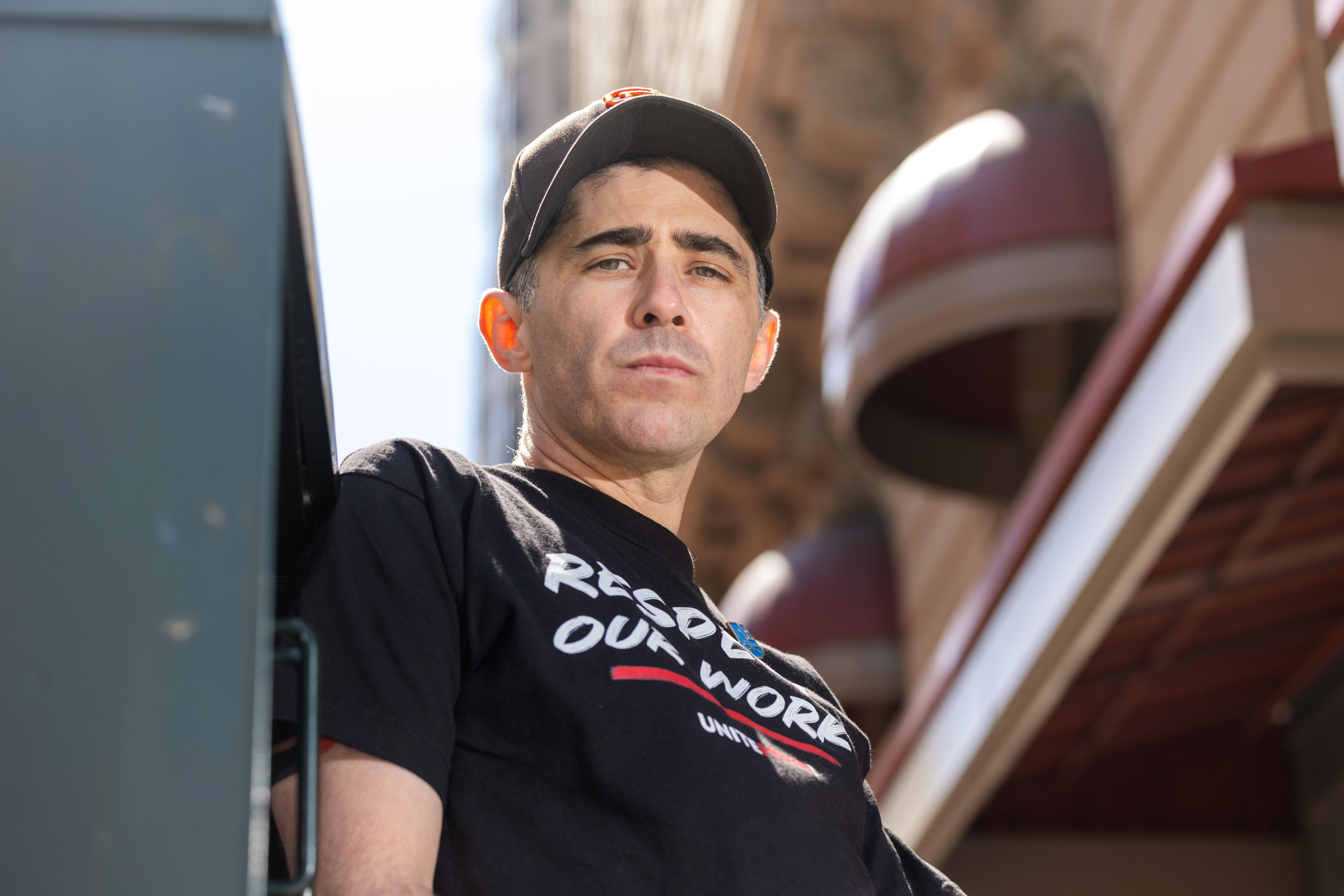 A person in a black cap and black T-shirt with the message &quot;RESPECT OUR WORK&quot; stands outdoors, looking forward confidently, with buildings in the background.