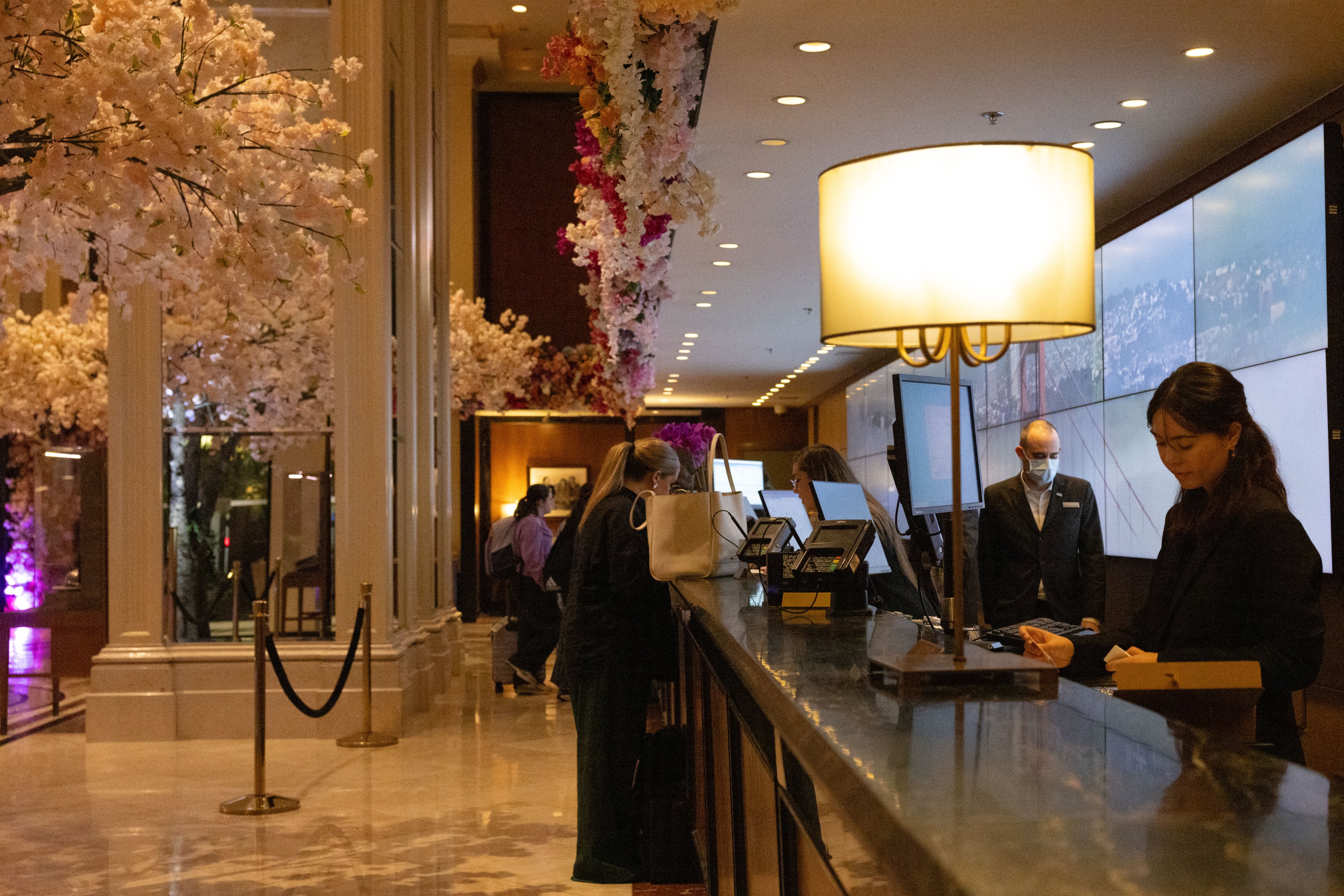 People are standing at a hotel reception desk, with pink floral decorations above. The area is warmly lit, featuring a lady with a tablet, and a man in a suit wearing a mask.