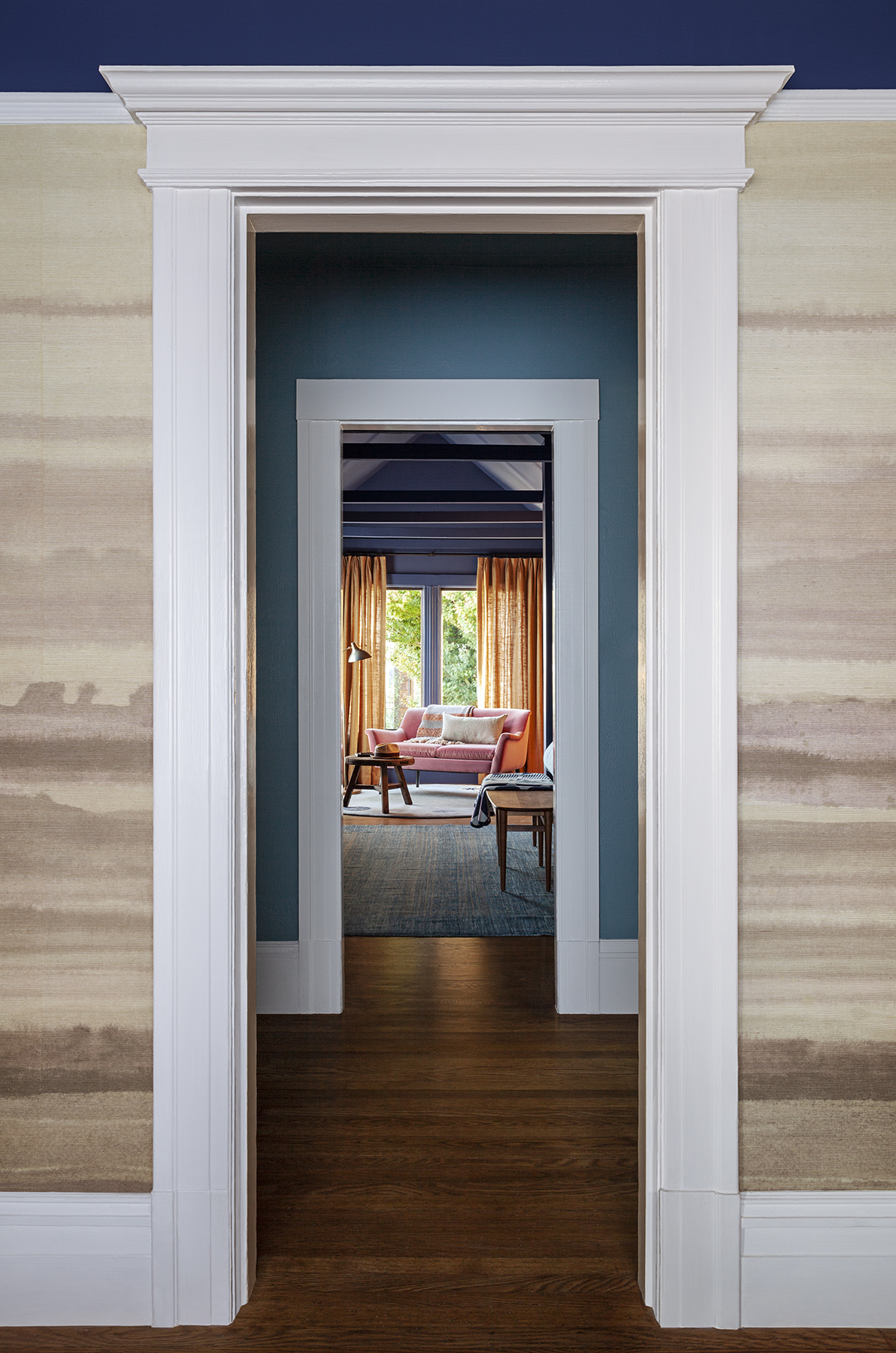 Through three aligned doorways, you see a cozy room with a pink sofa, wooden floors, blue walls, beige curtains, a lamp, and a window showcasing greenery.