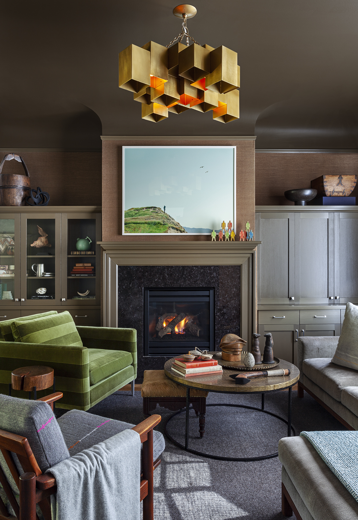 The image shows a cozy living room with a green armchair, a round coffee table, a fireplace, and modern shelving. A gold geometric chandelier hangs from the ceiling.