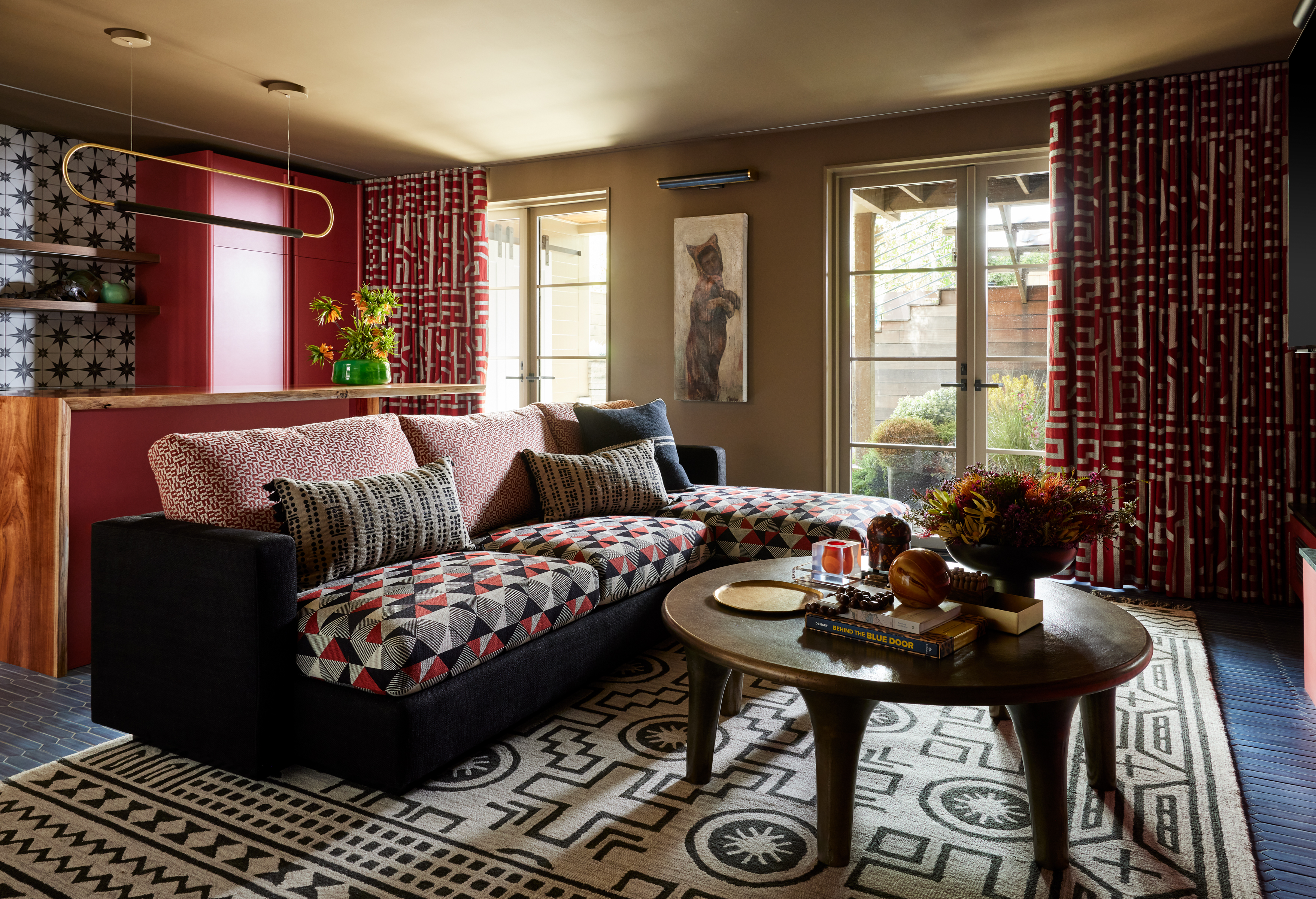 A cozy living room features a patterned sectional sofa, a round coffee table with decor, geometric carpet, red curtains, large windows, and vibrant wall art.