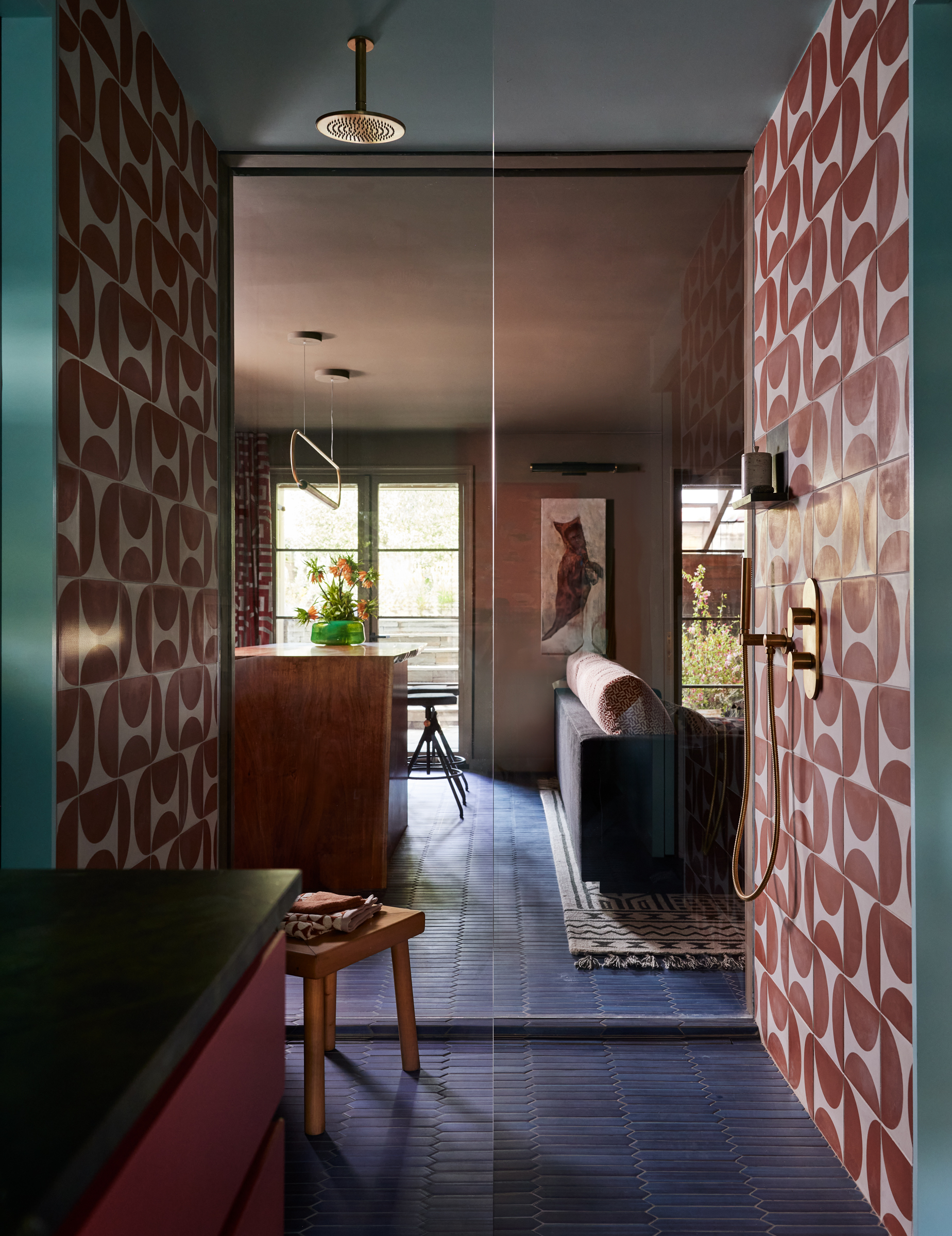 A modern bathroom with blue floor tiles and walls adorned with red and white geometric patterns opens into a stylish living area with a couch, plants, and artwork.