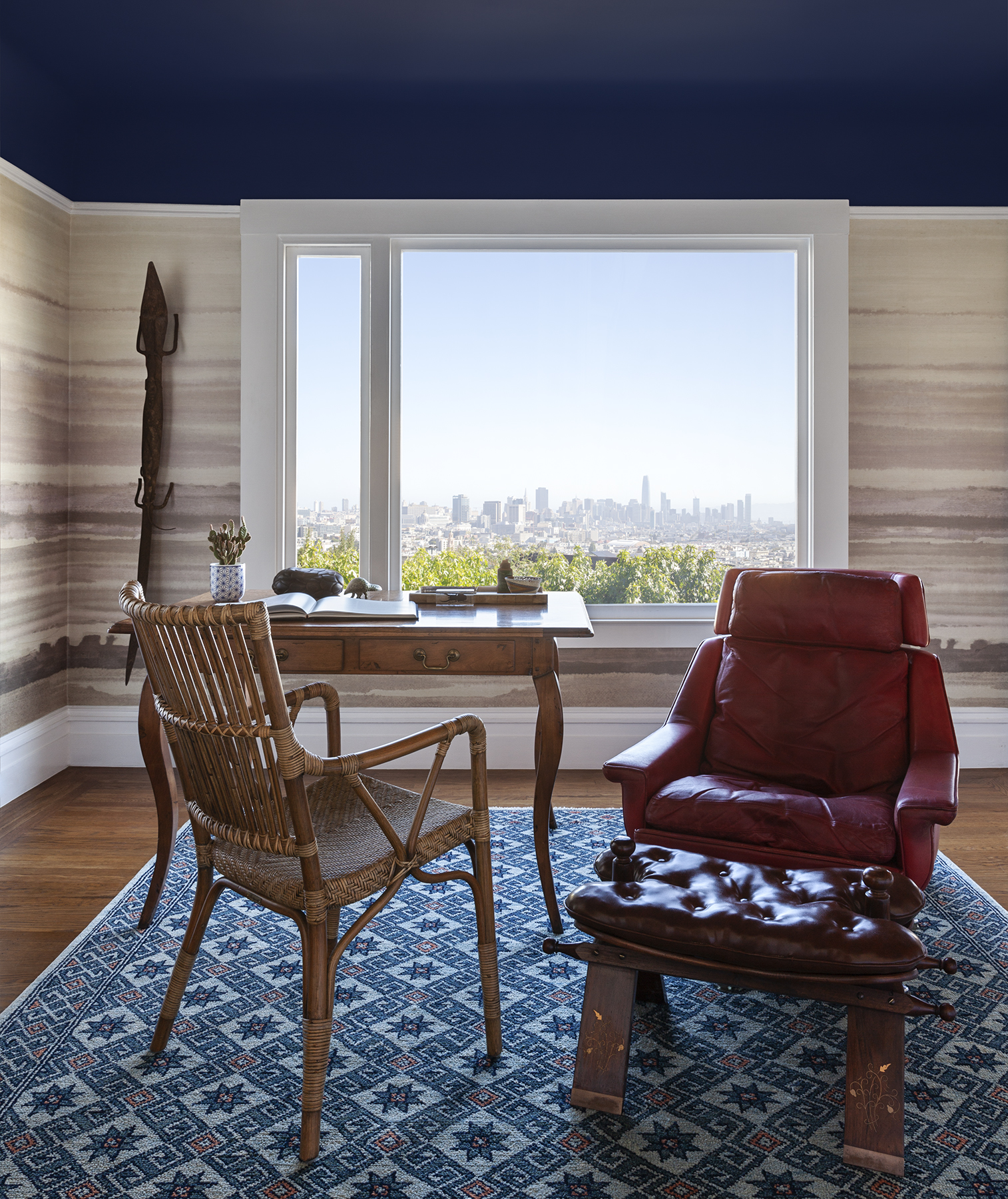 A room with a wooden desk, a wicker chair, and a red leather lounge chair faces a large window overlooking a city skyline. There's a blue patterned rug on the floor.