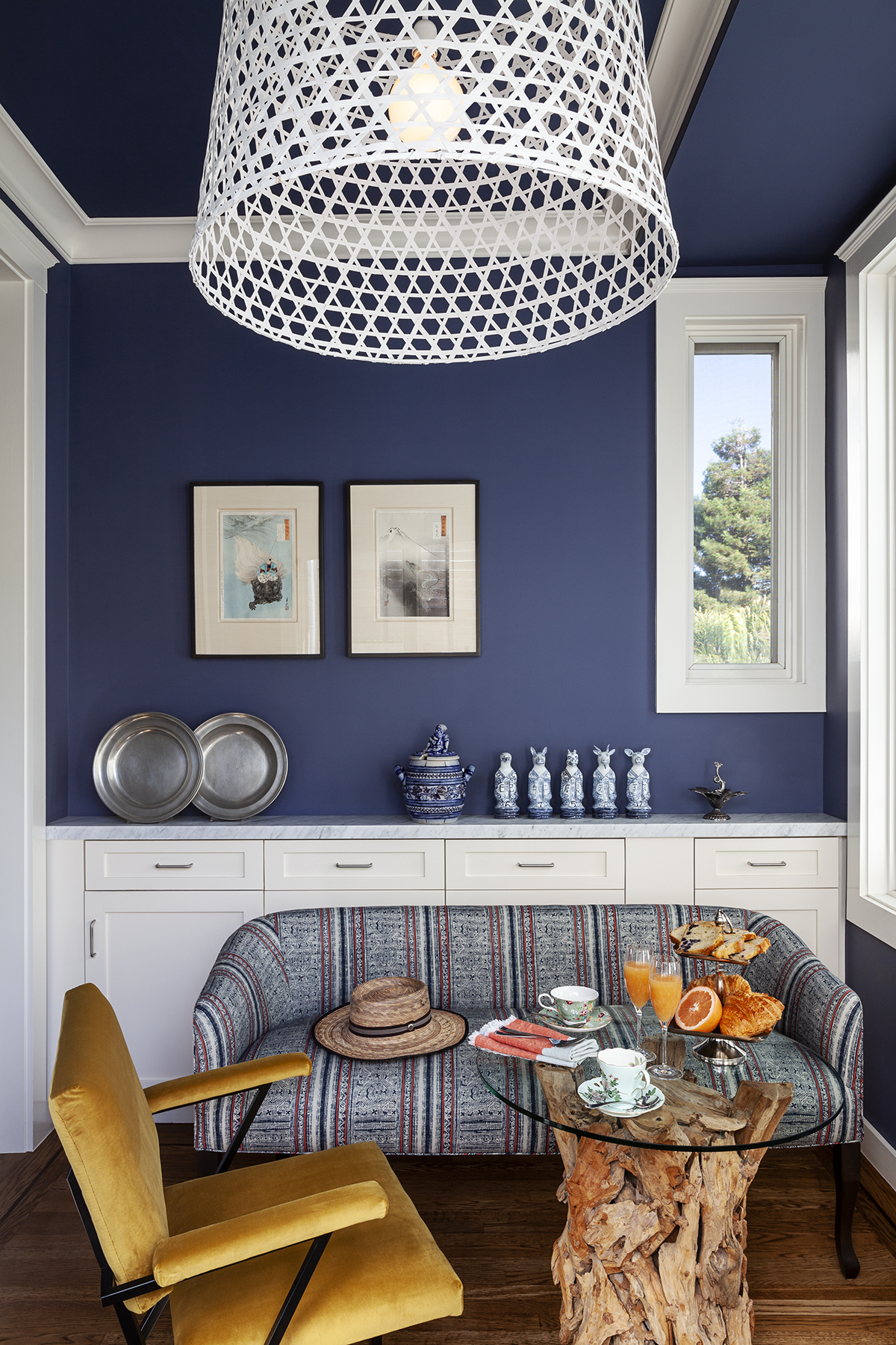 A cozy room with blue walls features eclectic decor, a yellow chair, a patterned couch, and a rustic table with breakfast items. A large white woven light hangs overhead.