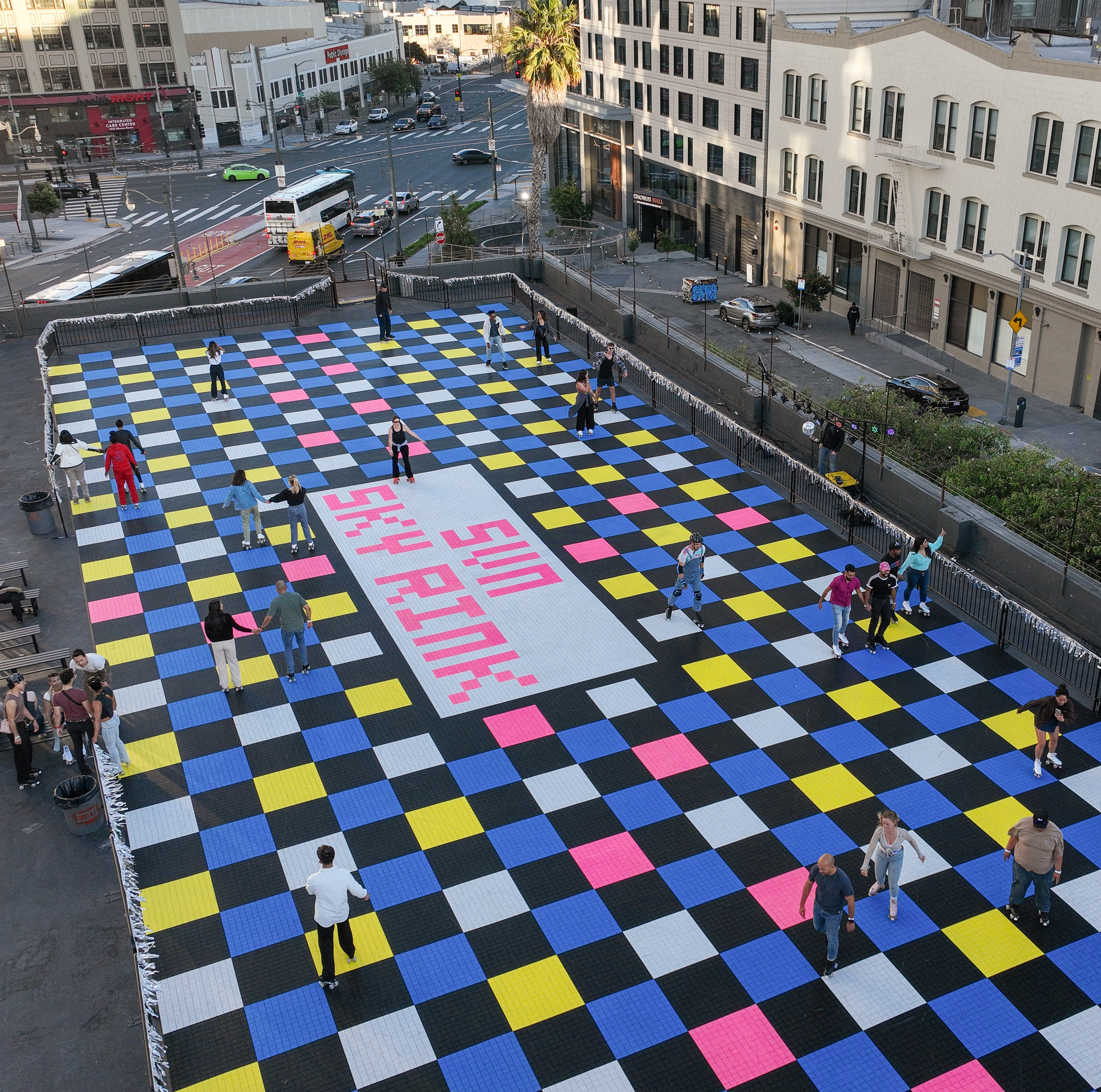 People skate on a large, checkered rink with black, blue, white, yellow, and pink squares in a city. A white rectangular area with &quot;SKY RINK&quot; in pink text is visible.