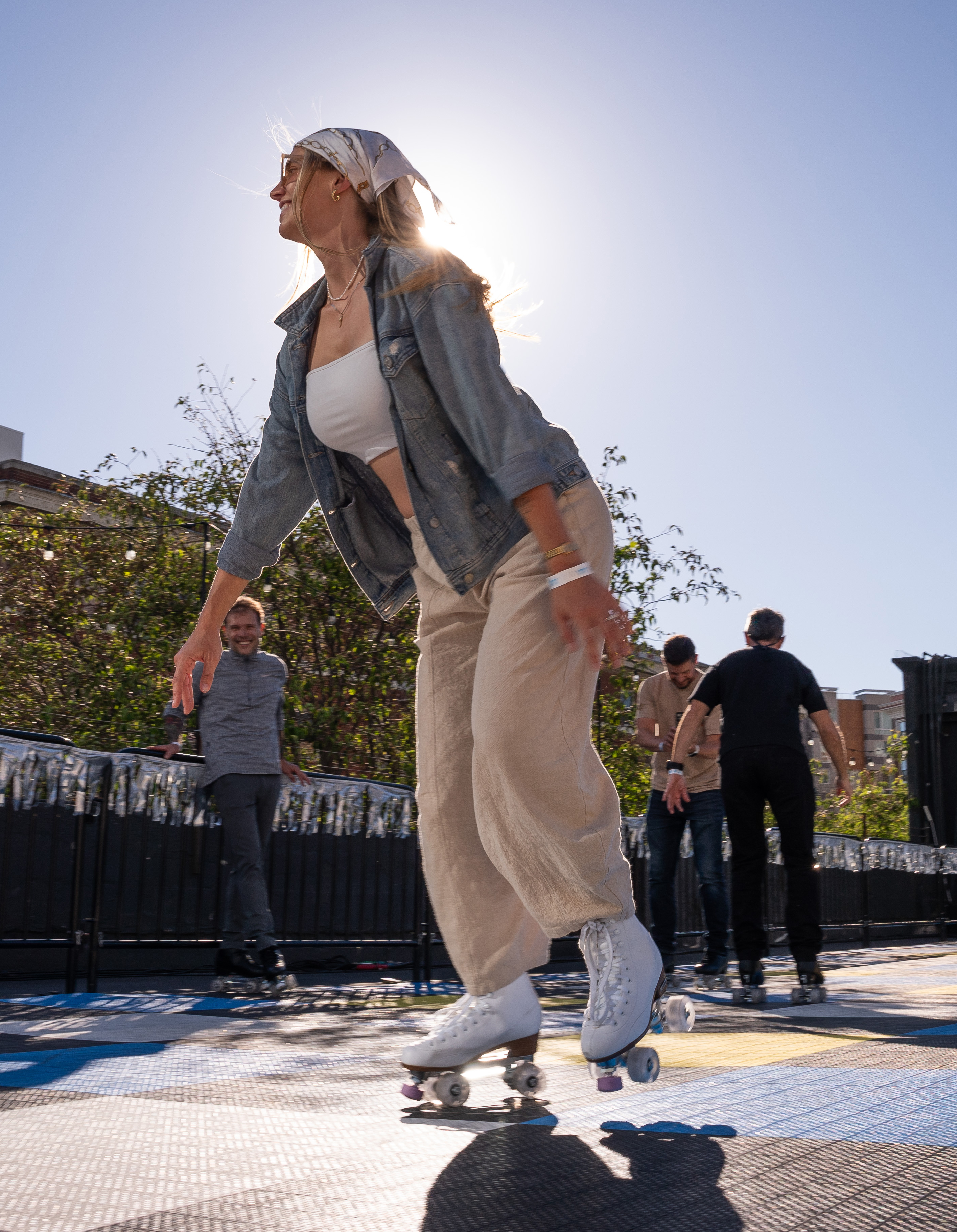 A person in a denim jacket and beige pants roller-skates in an outdoor rink, the sun shining behind them. Other skaters and trees are visible in the background.