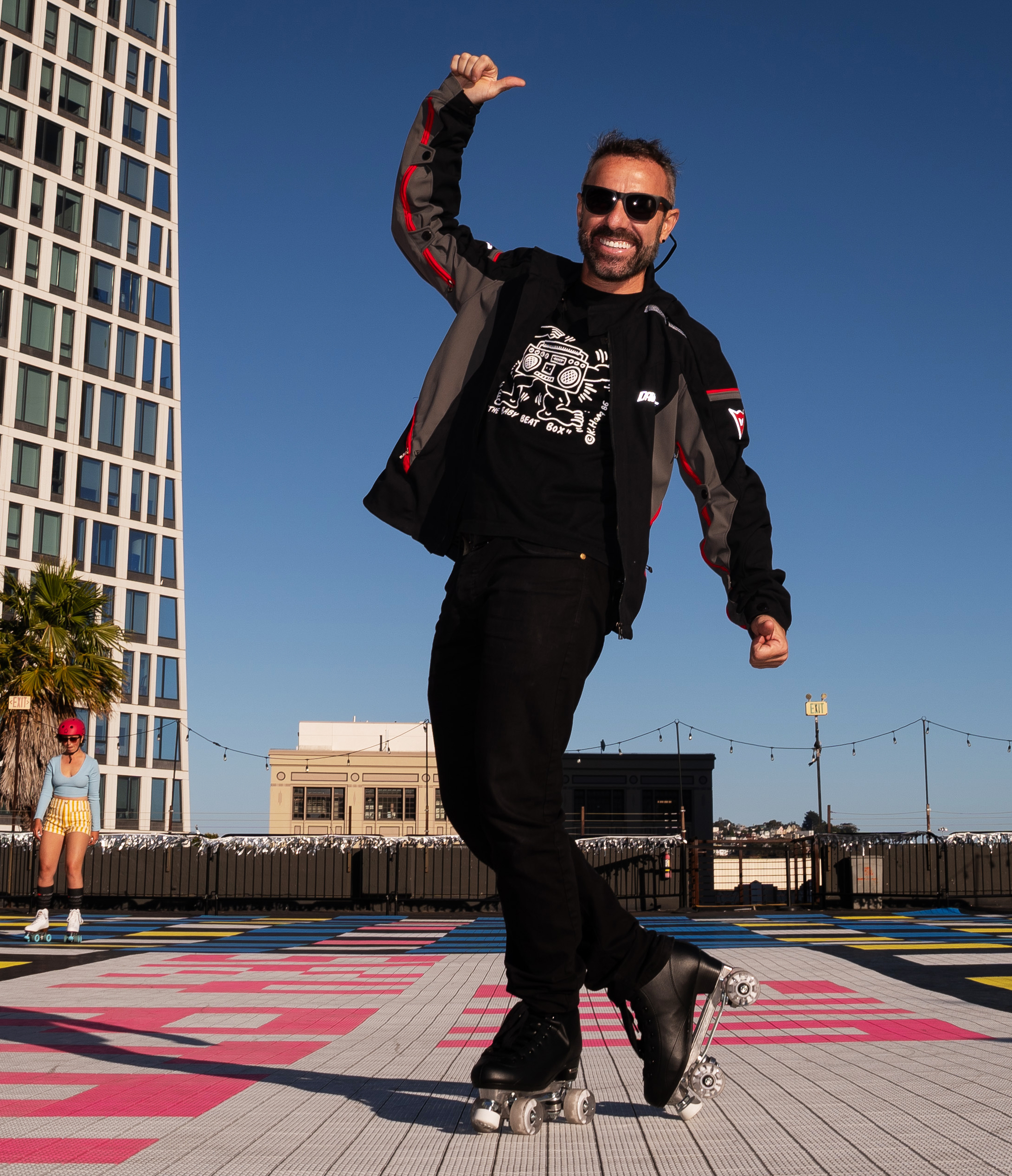 A man in roller skates, wearing a black outfit and sunglasses, poses energetically on a colorful rooftop with modern buildings and a woman skating in the background.