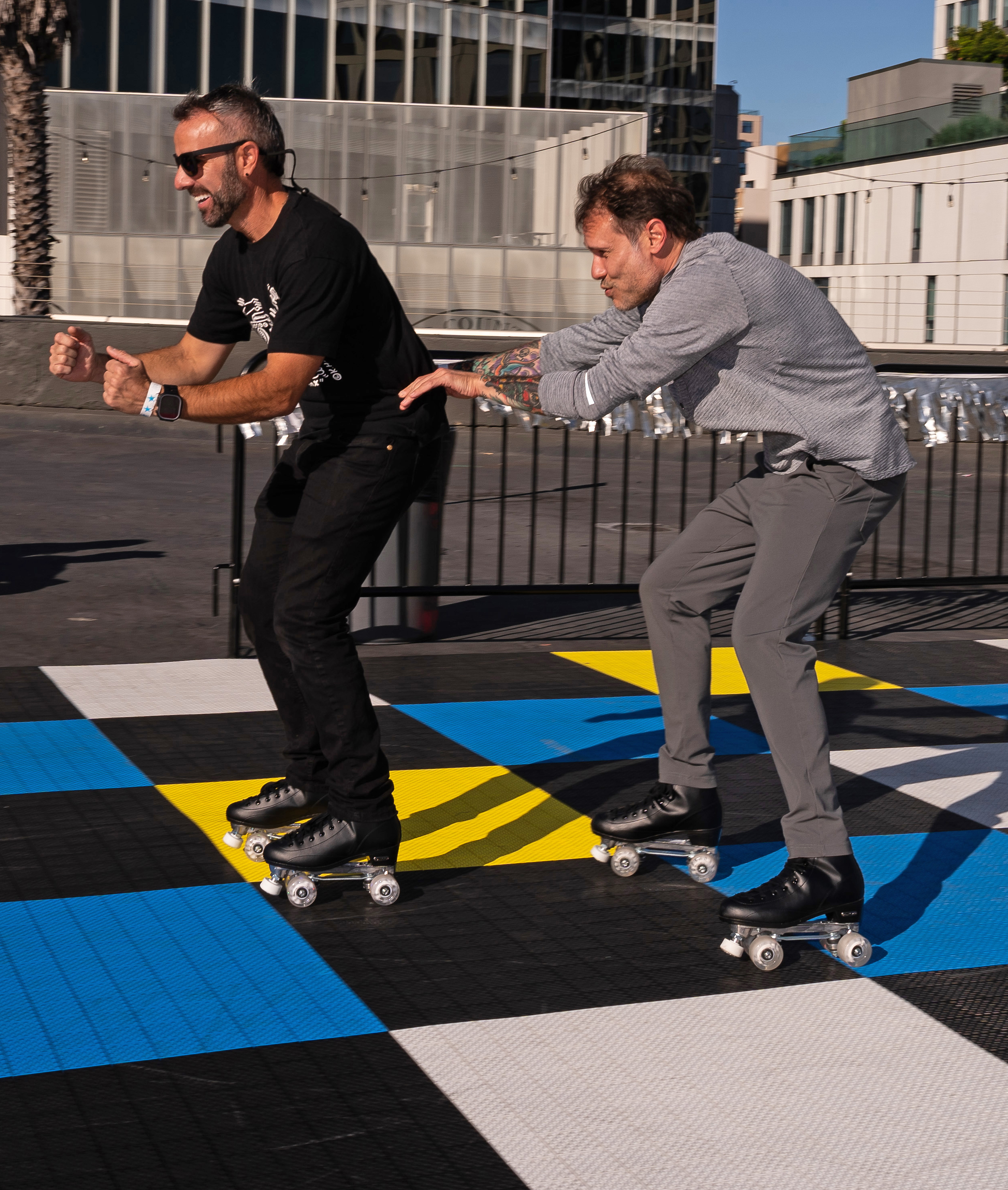 Two men are roller skating on a colorful checkered surface outdoors. The man in black is ahead and laughing, while the man in gray follows closely, pushing his back.