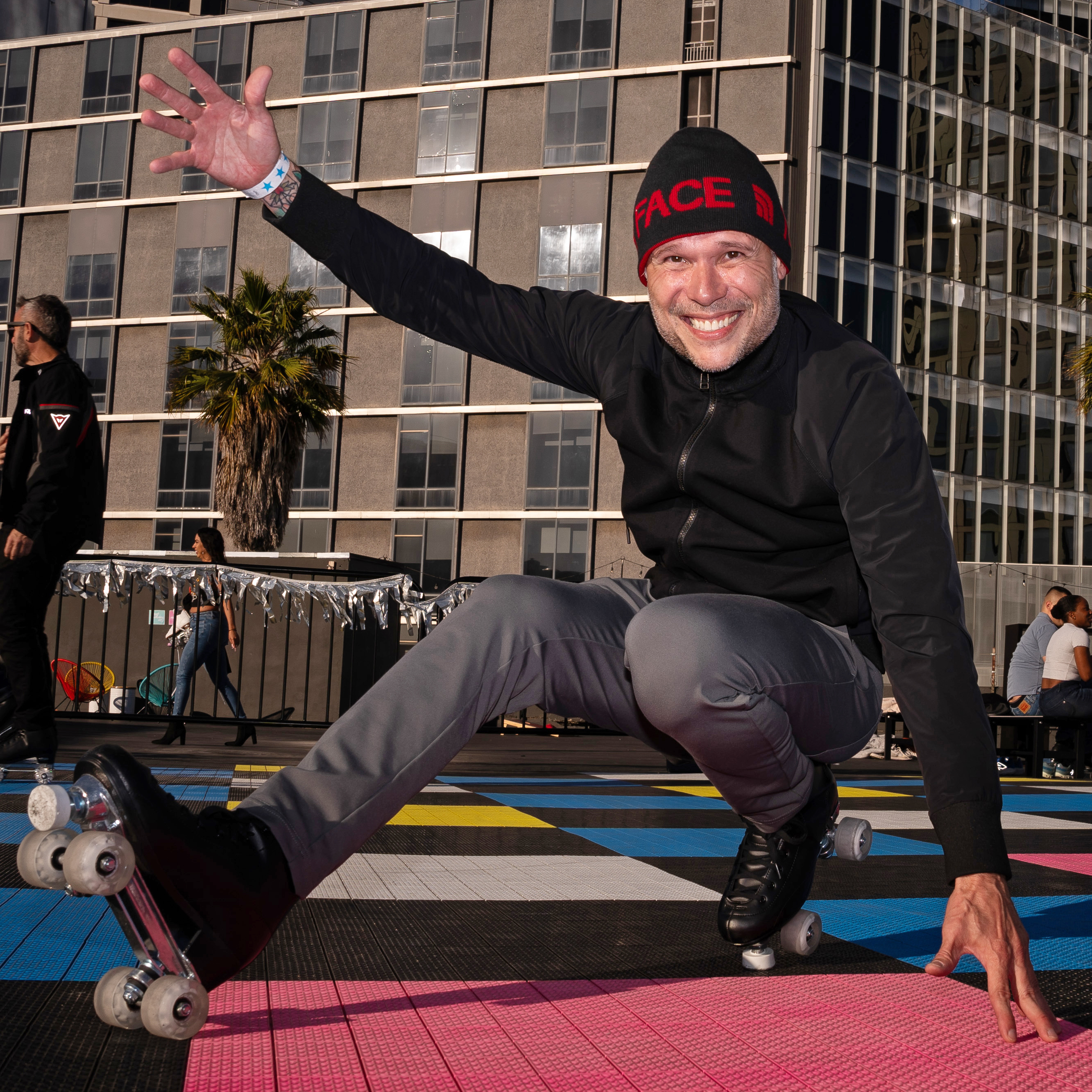 A man in a black beanie and jacket is smiling and roller skating on a colorful checkered floor, with a modern building and other people in the background.