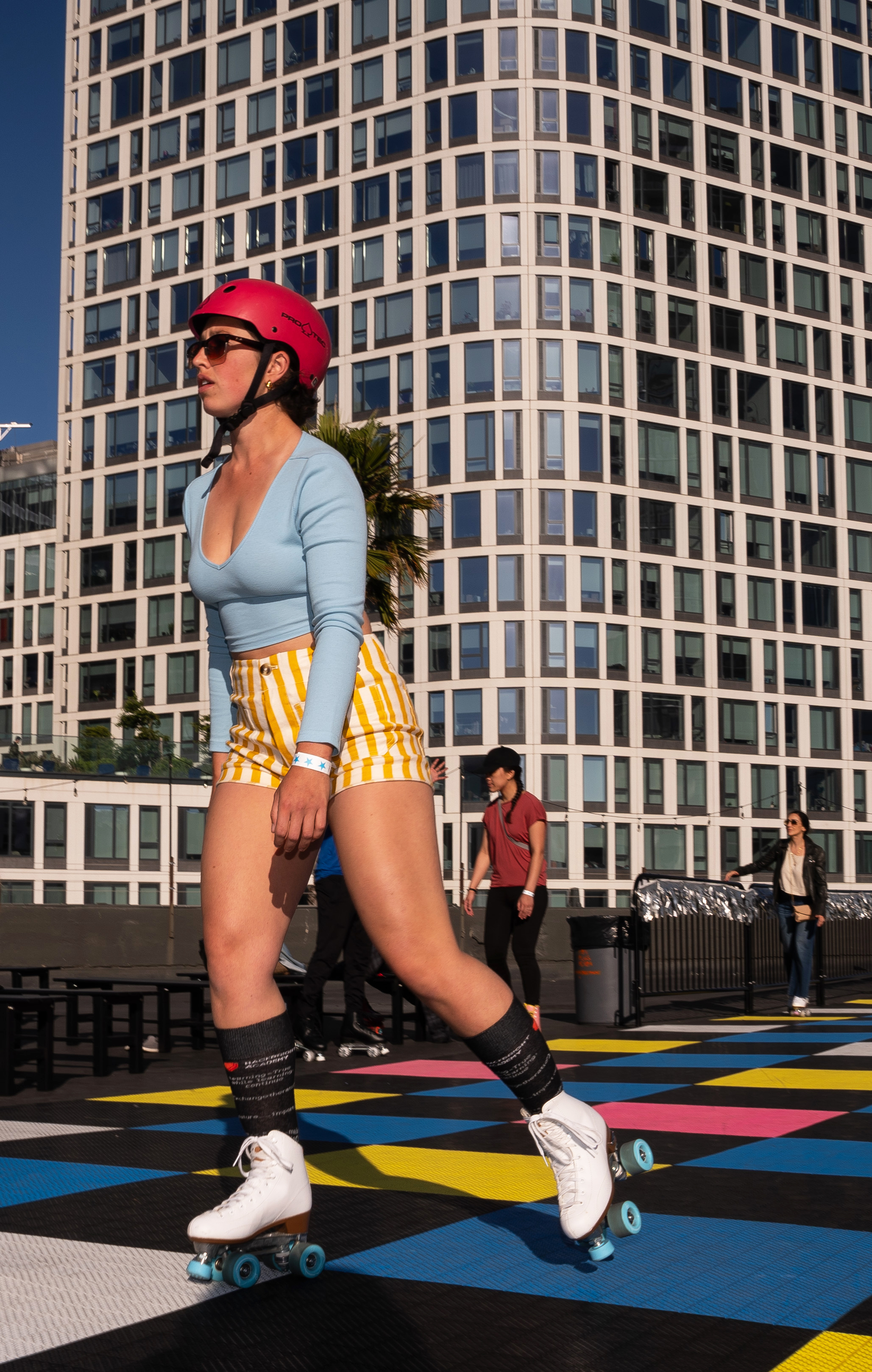 A woman wearing a red helmet, blue top, and yellow striped shorts is roller skating on a colorful surface in front of a large modern building.