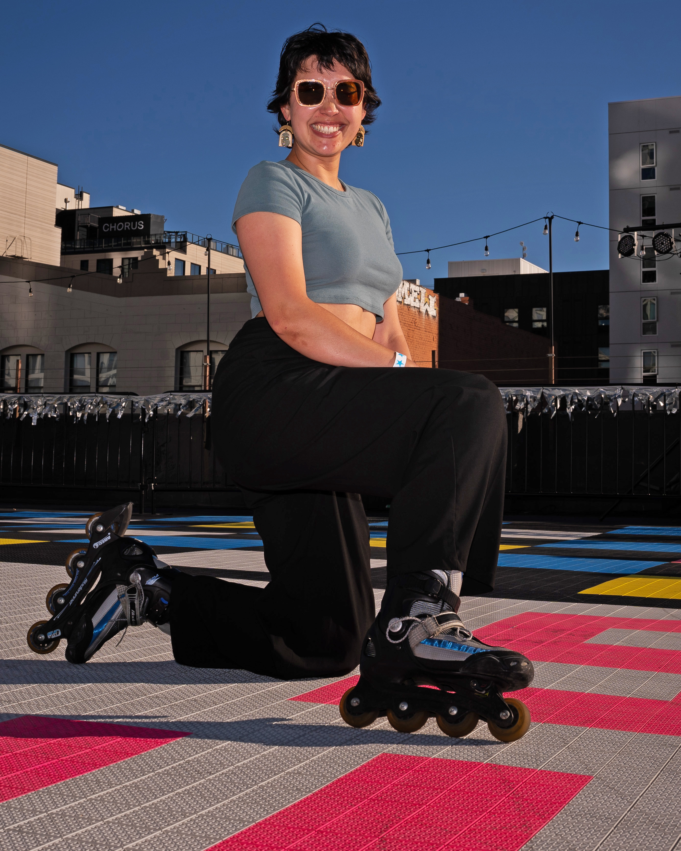 A person in sunglasses and a cropped shirt is smiling at the camera while kneeling on rollerblades on a colorful outdoor skating rink with buildings in the background.