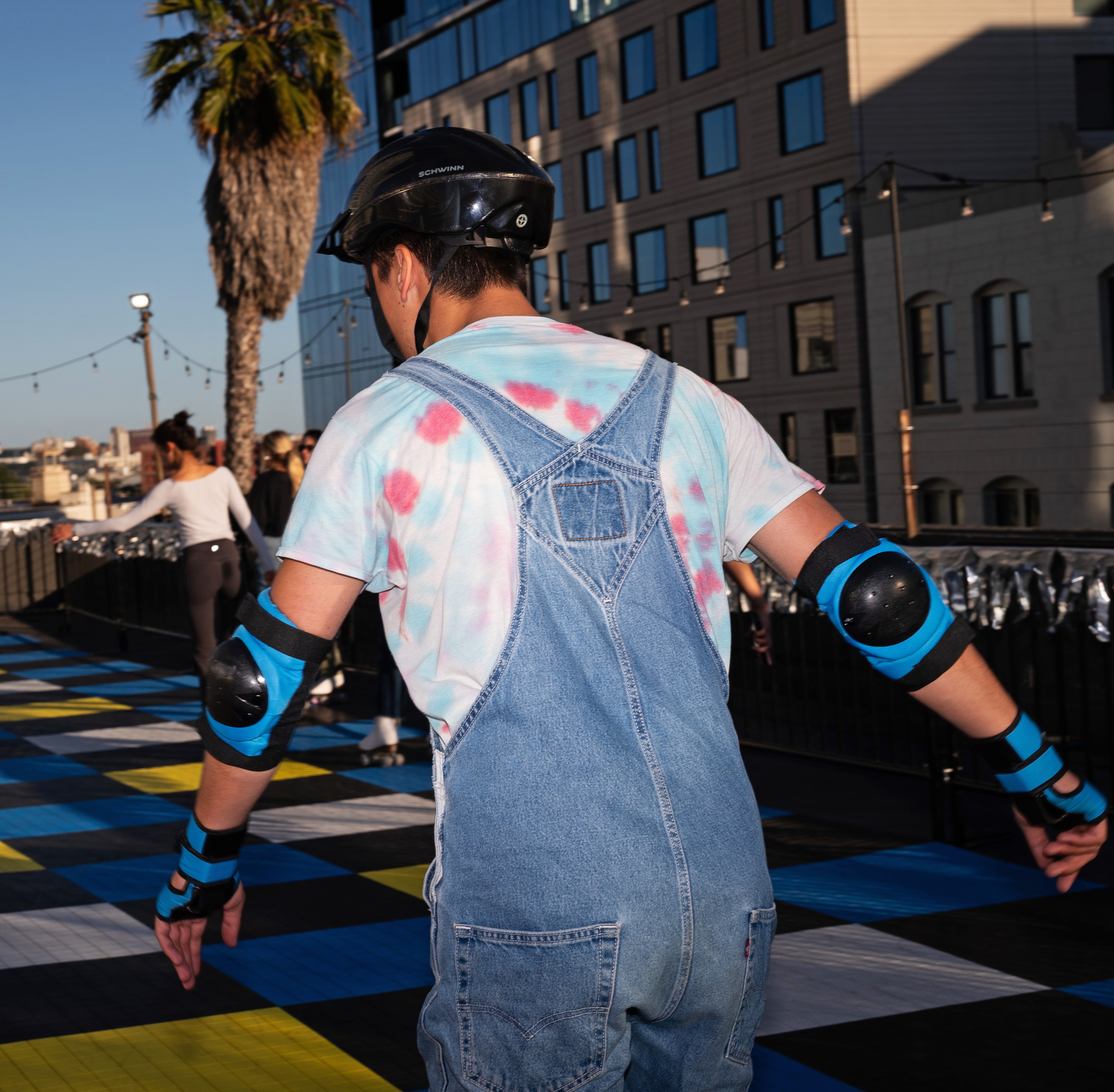 A person wearing a tie-dye shirt and denim overalls is roller skating with protective gear. They're on a colorful checkered path, with buildings and a palm tree in the background.