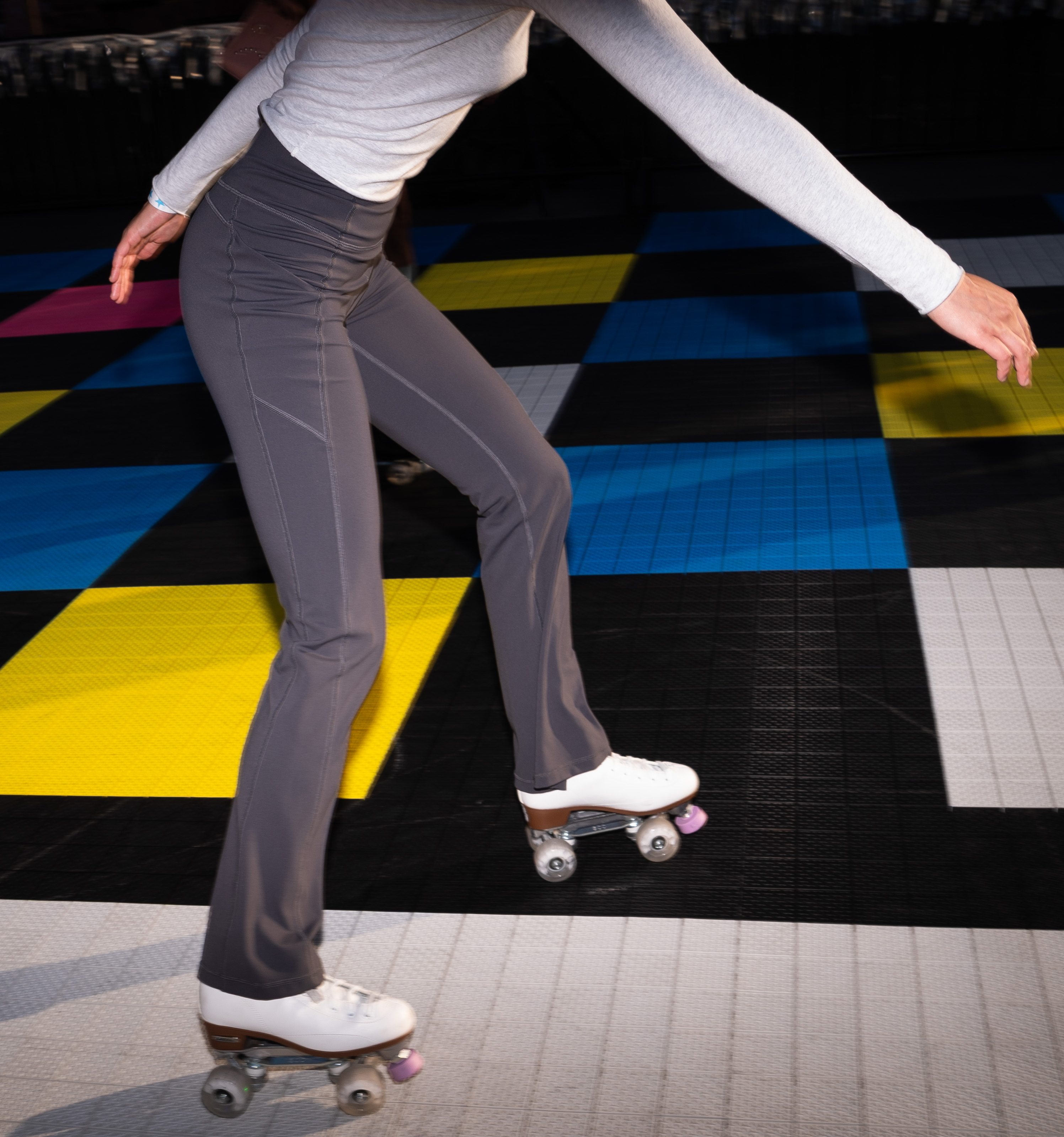 A person in gray pants and a light long-sleeve top is roller skating on a colorful, checkered floor with yellow, blue, black, and white tiles.