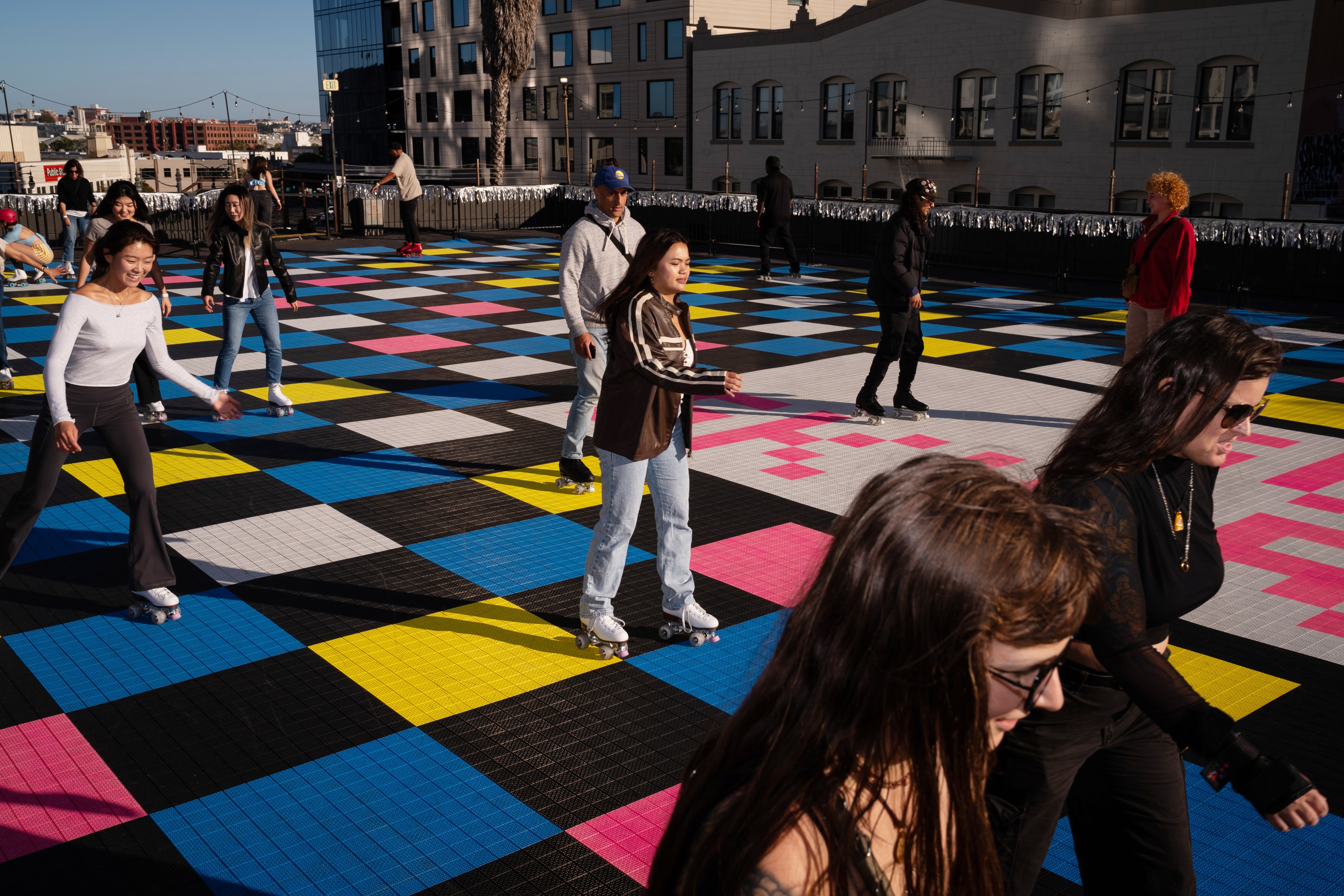 A group of people are roller skating on a brightly colored, checkered outdoor rink, with a city skyline and buildings in the background. Elements include bright sunlight and various skaters.