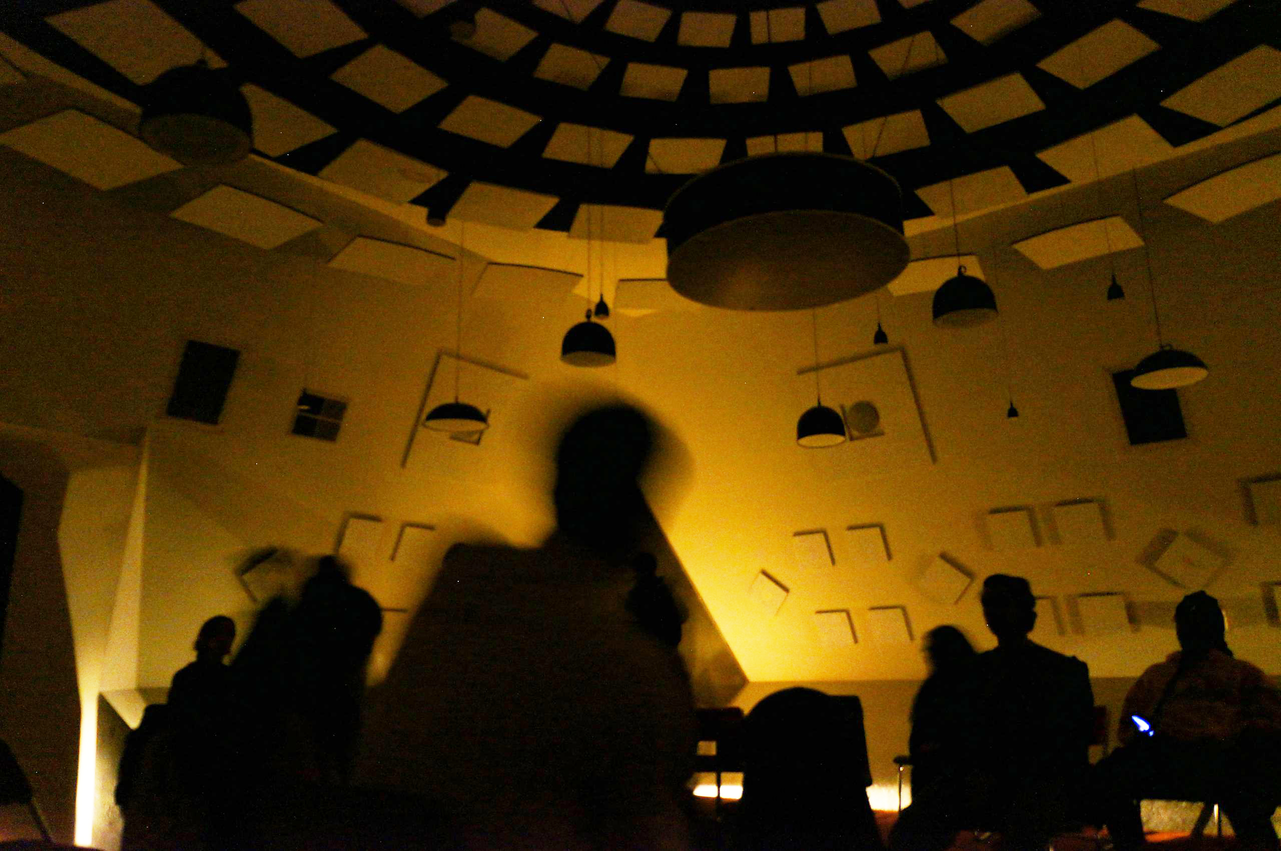 People are silhouetted against a warmly lit, patterned ceiling with hanging lights, giving a moody ambiance. The abstract shapes on the walls add to the artistic feel.