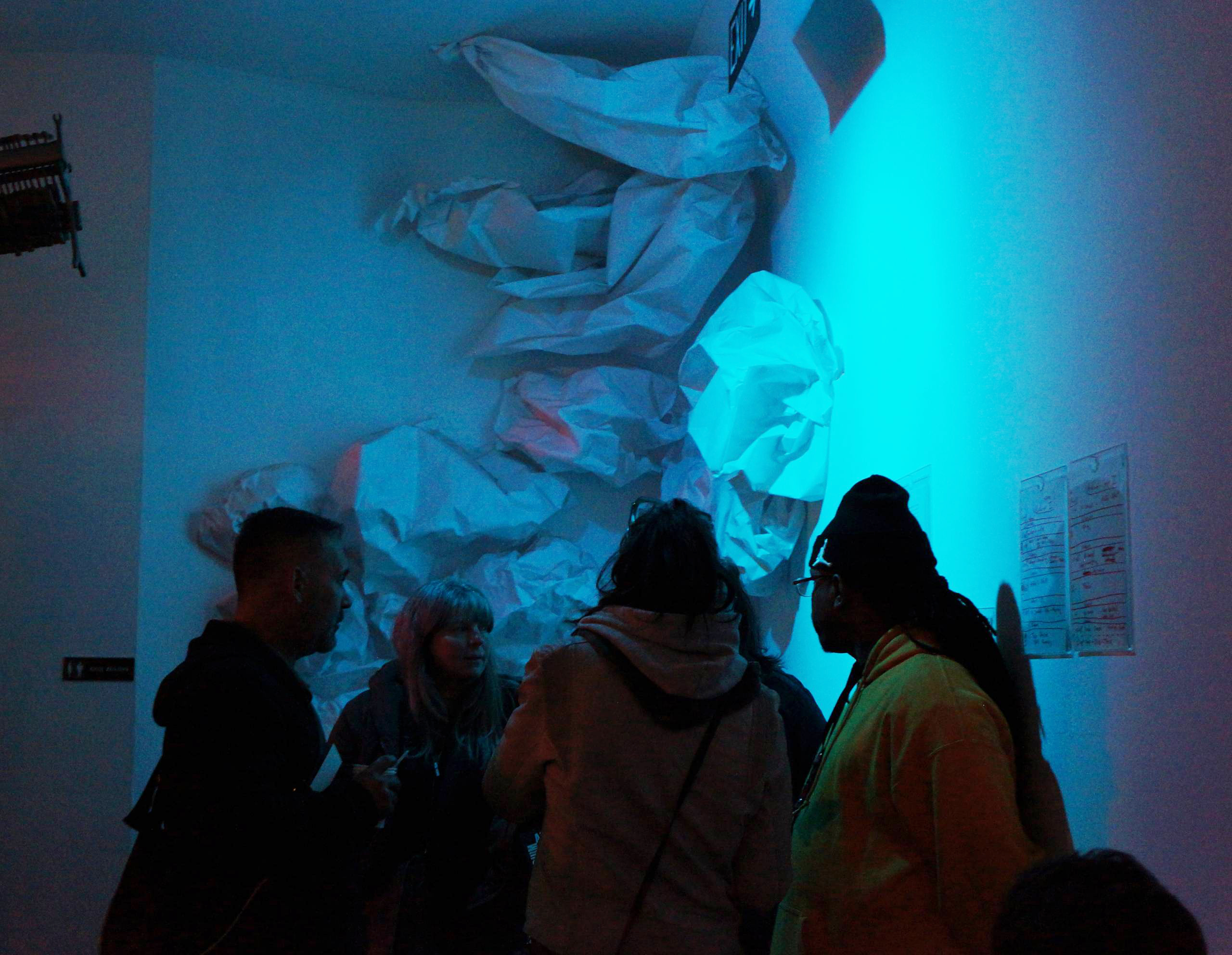 In blue lighting, four people gather near a wall decorated with large, crumpled white paper formations. Various objects and documents are affixed to the walls.