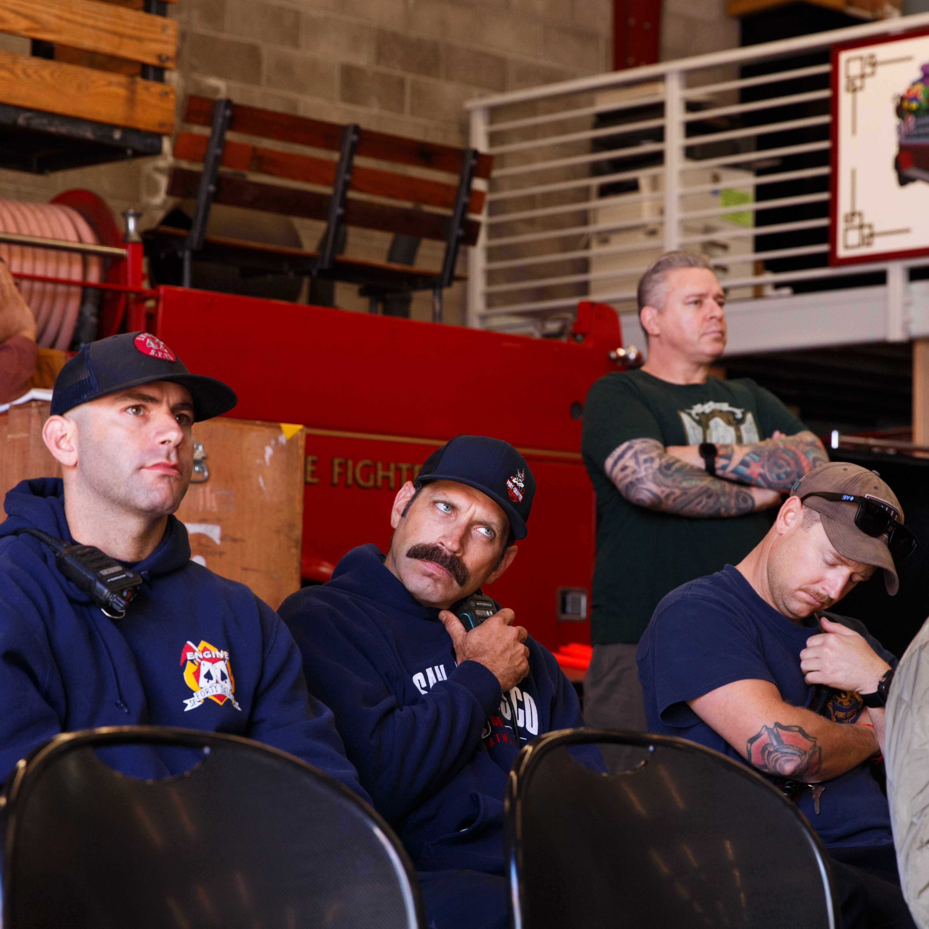 Several people, including uniformed firefighters, sit and listen attentively in a firehouse. A red fire engine and a &quot;San Francisco&quot; sign are visible in the background.