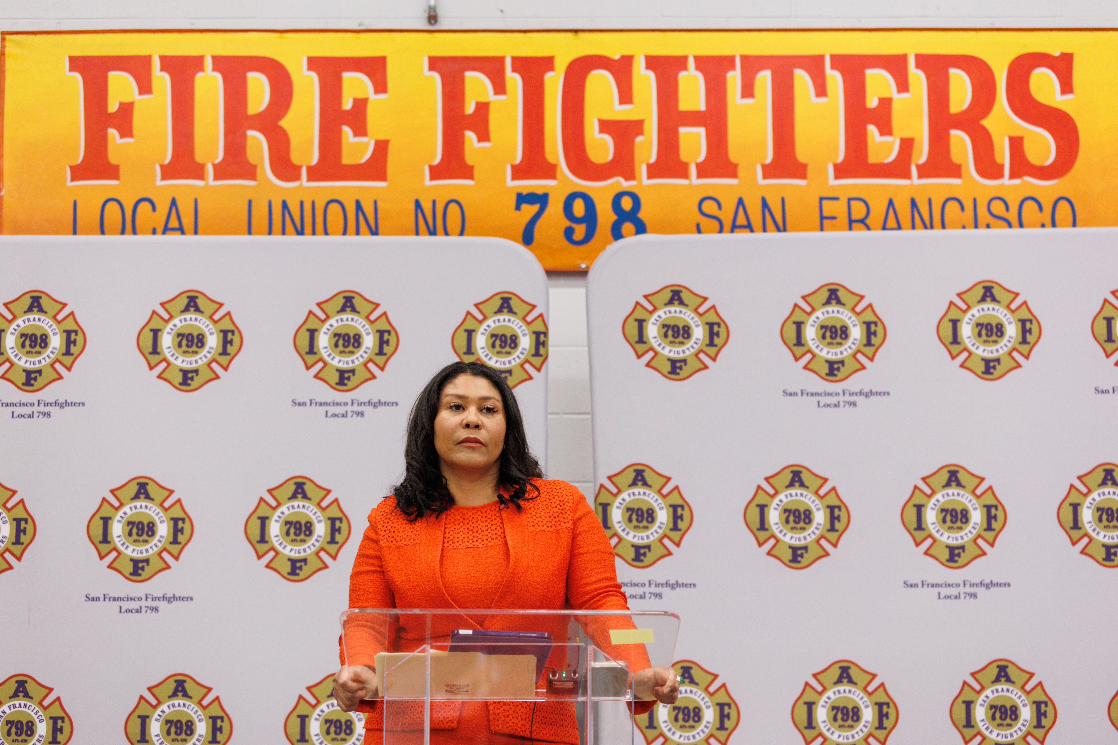 A person in an orange outfit stands at a podium with &quot;San Francisco Firefighters Local 798&quot; logos behind and a &quot;Fire Fighters Local Union No. 798 San Francisco&quot; sign above.