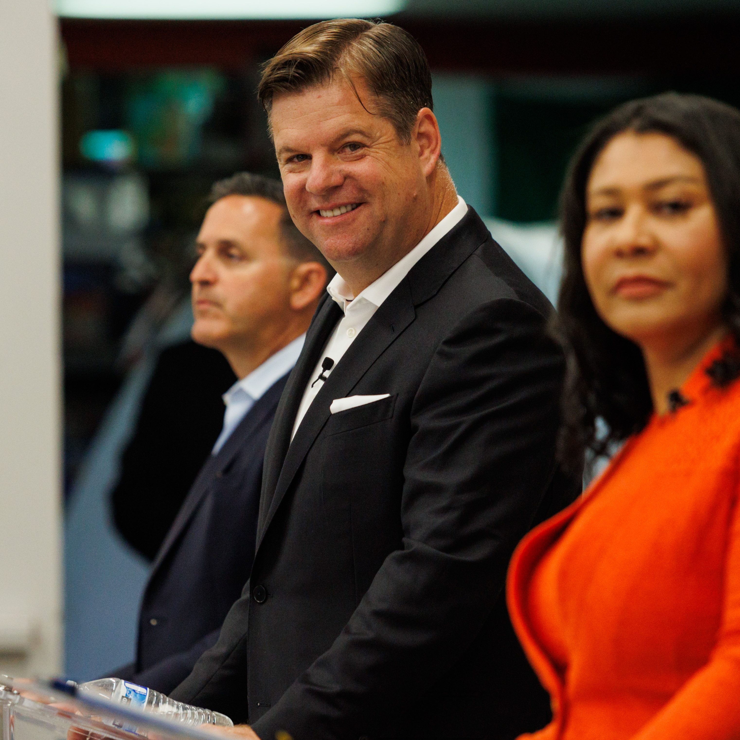 Three people in business attire stand side by side. The central figure is smiling, while the others have neutral expressions. The man in the middle looks directly at the camera.