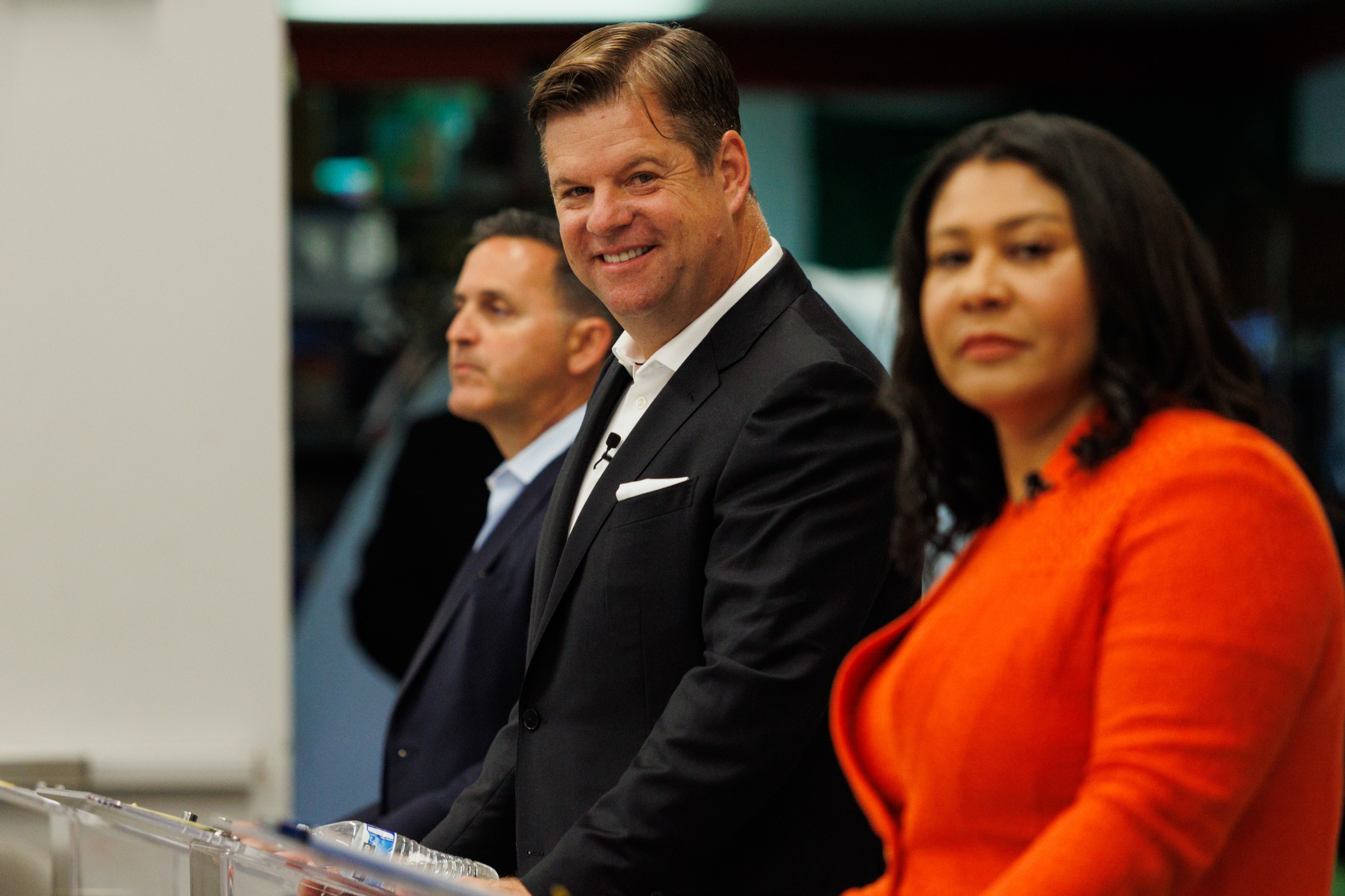 Three people in business attire stand side by side. The central figure is smiling, while the others have neutral expressions. The man in the middle looks directly at the camera.