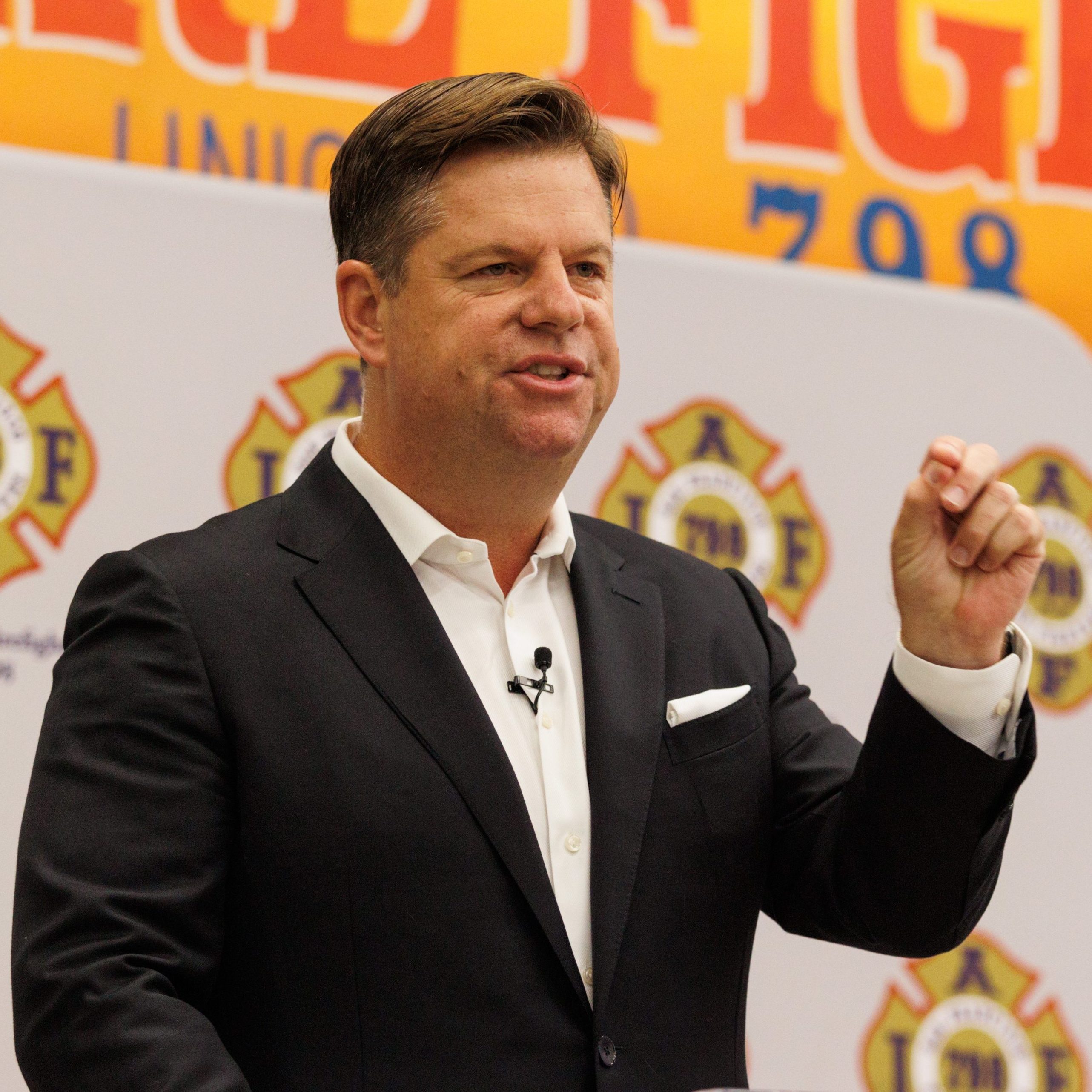 A man in a suit speaks passionately at a podium, with a large, bright &quot;Fire Fighters Local 798 San Francisco&quot; sign and firefighter logos behind him.