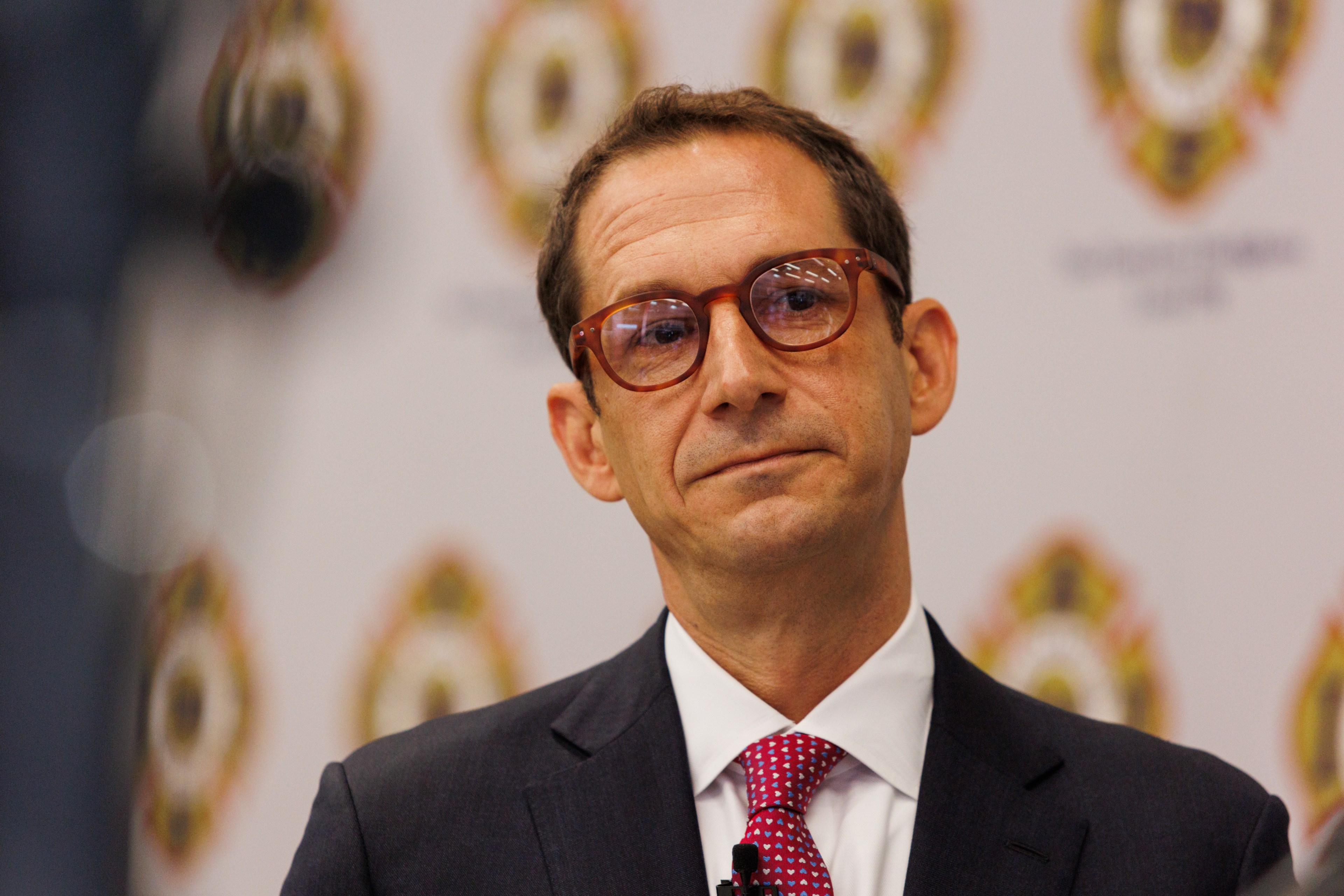 A man with glasses, wearing a dark suit and a pink tie, is standing against a backdrop with a repeating official emblem pattern.