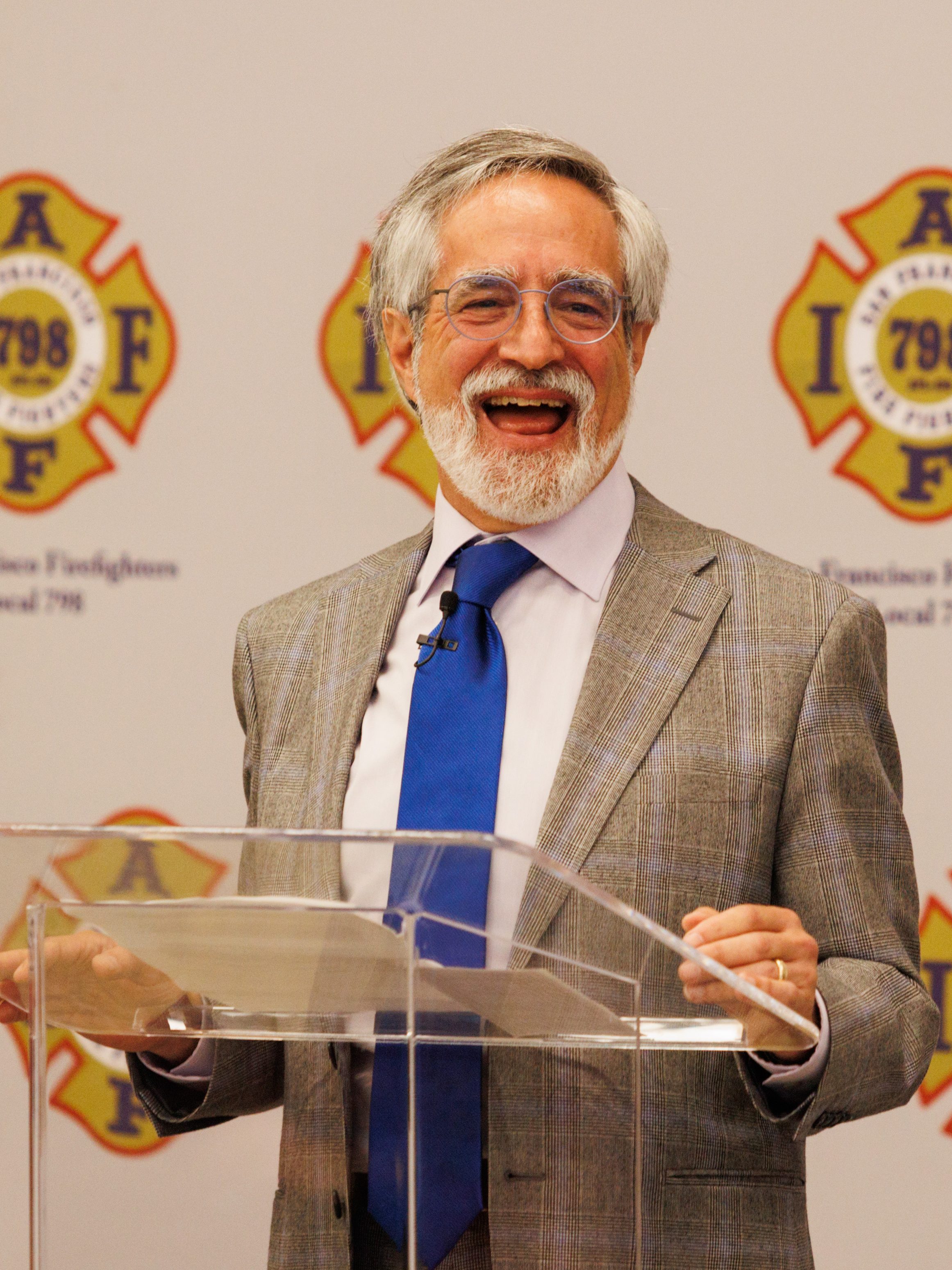 A bearded man holding a mic, in a gray suit and blue tie, is smiling widely at a clear podium with firefighter emblems in the background.