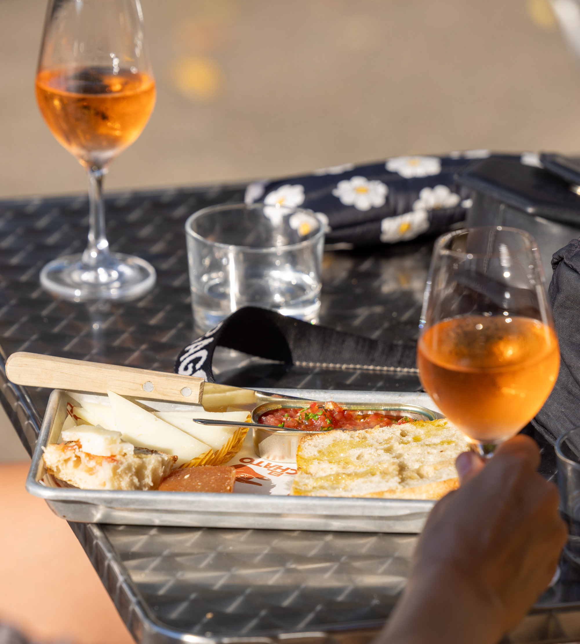 Two glasses of rose wine, an empty glass, a tray with cheese, bread, and a tomato-based dip on a metal table with a floral-patterned item in the background.