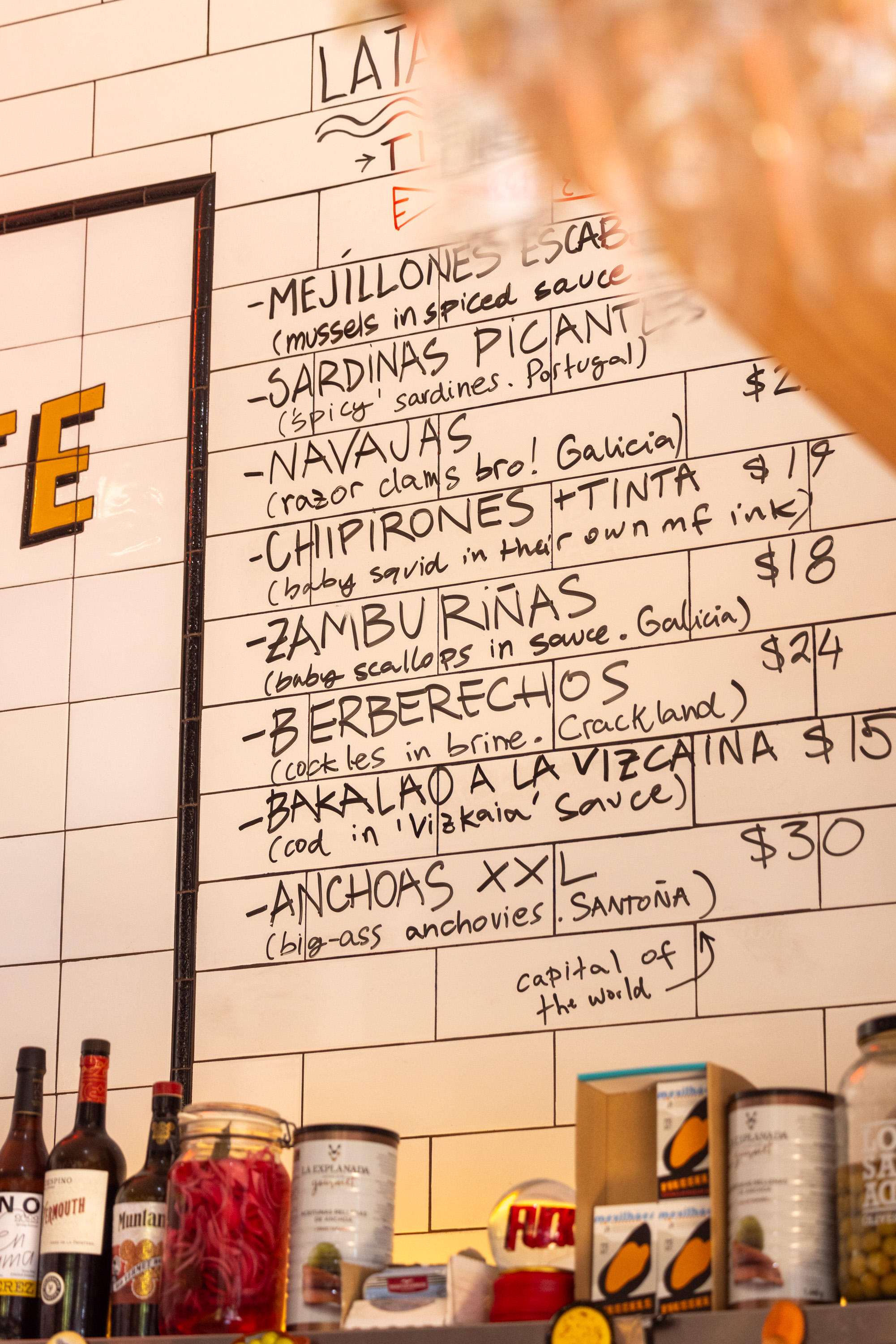 The image shows a handwritten menu on a white tiled wall listing various seafood dishes with prices. Bottles and jars are placed on a shelf below the menu.