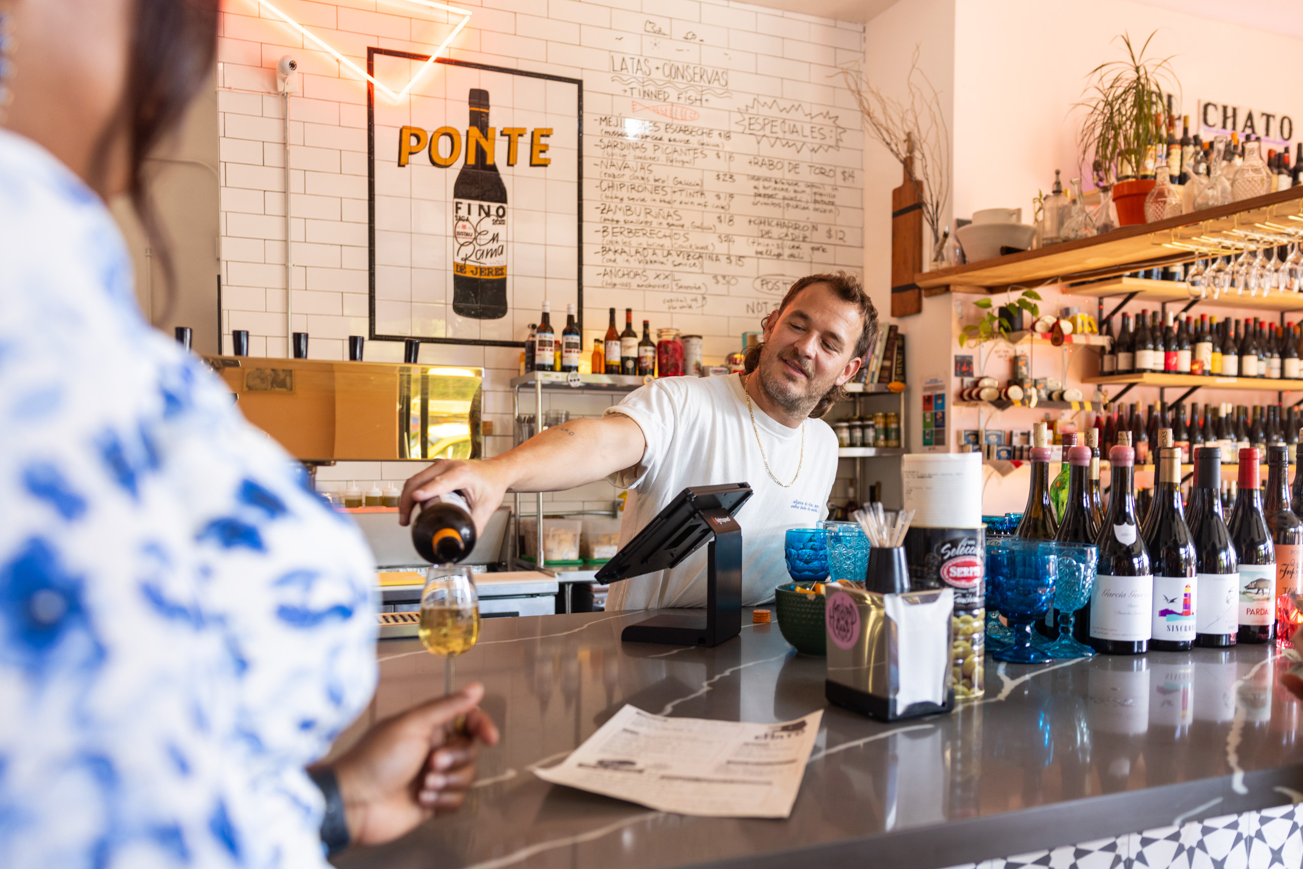 A man behind a bar pours a drink for a customer. The bar has a tiled counter, wine bottles, blue glasses, and a menu board on a white tiled wall.