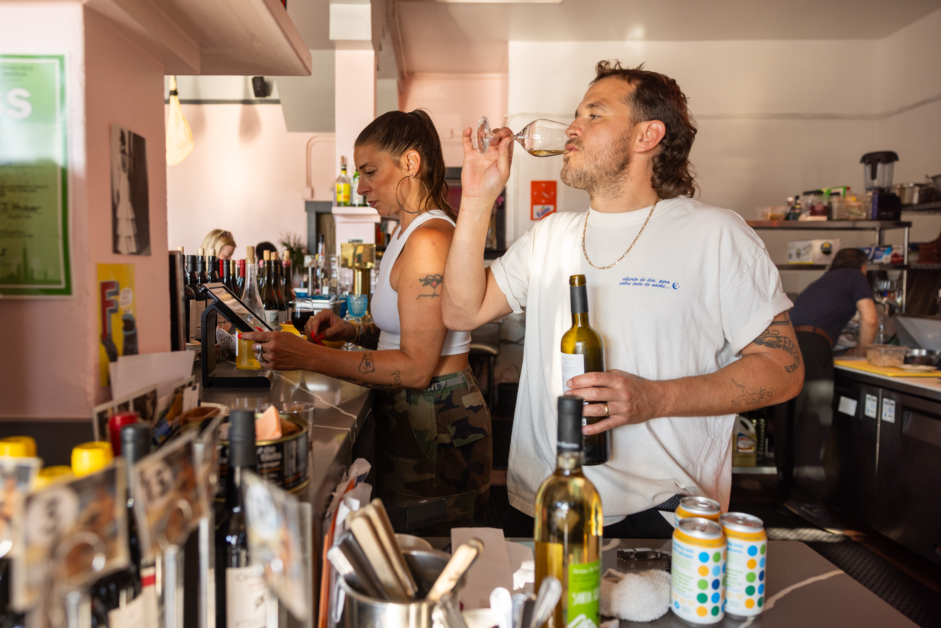 Two people are behind a busy bar; one is using a touchscreen while the other drinks from a wine glass, holding a wine bottle. The bar has various bottles and cans.