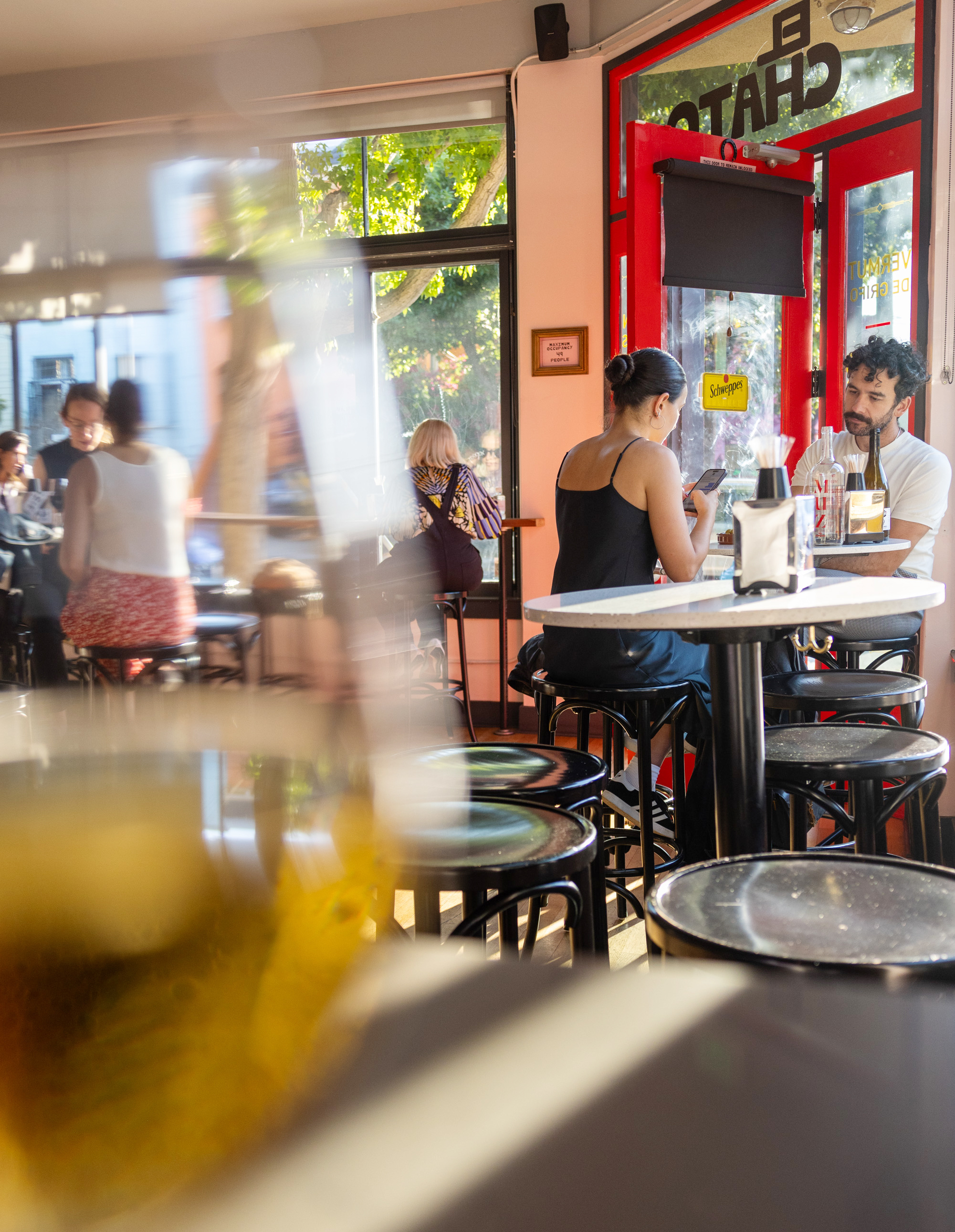 The image shows a cozy, sunlit café with several people sitting at tables and chatting. The focus is on two patrons near a window who are engrossed in their conversation. The outside greenery is visible through large windows.