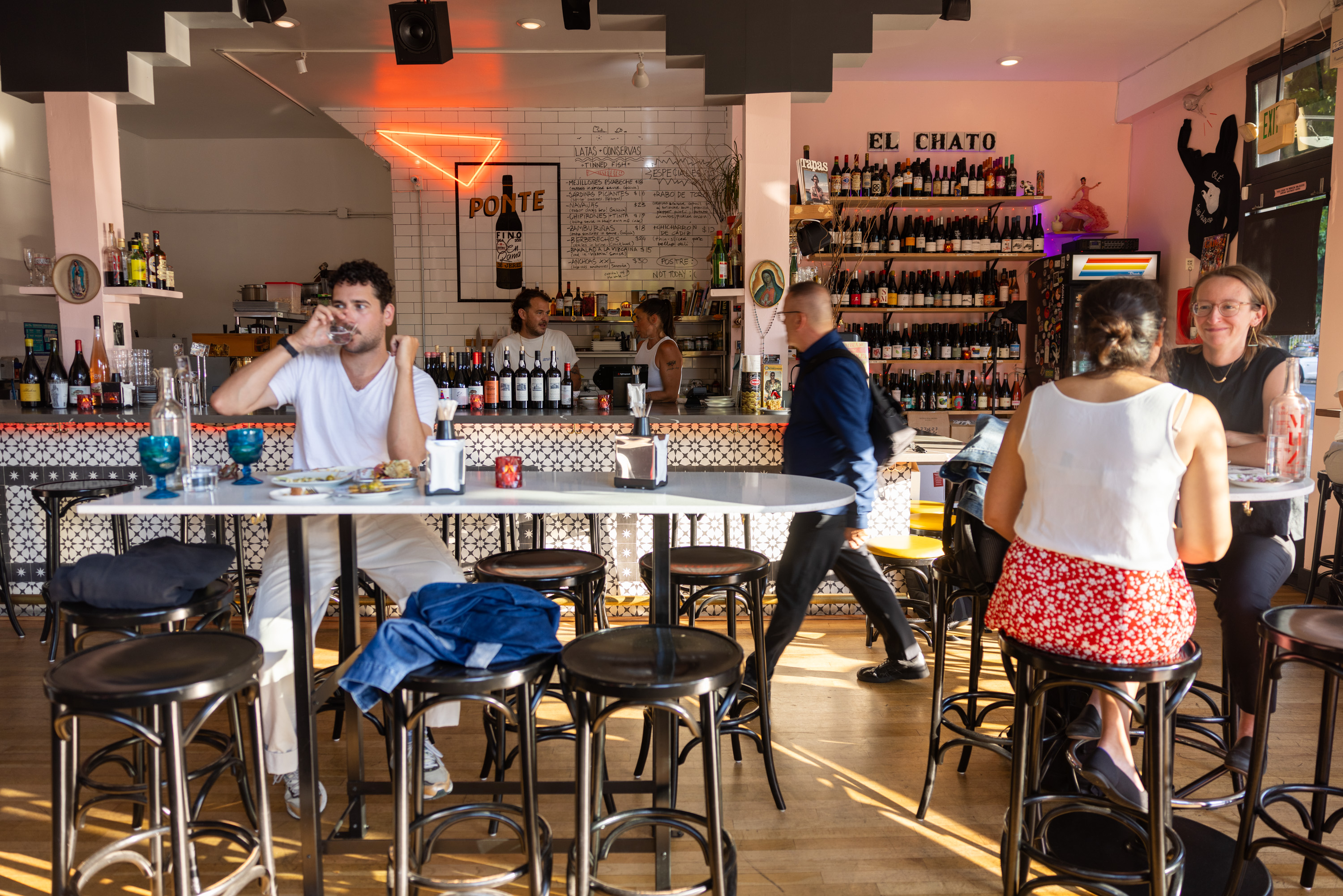 The image shows a vibrant bar scene with various people seated at tall tables, drinking and talking. Bottles line shelves behind the counter, with bartenders preparing drinks.