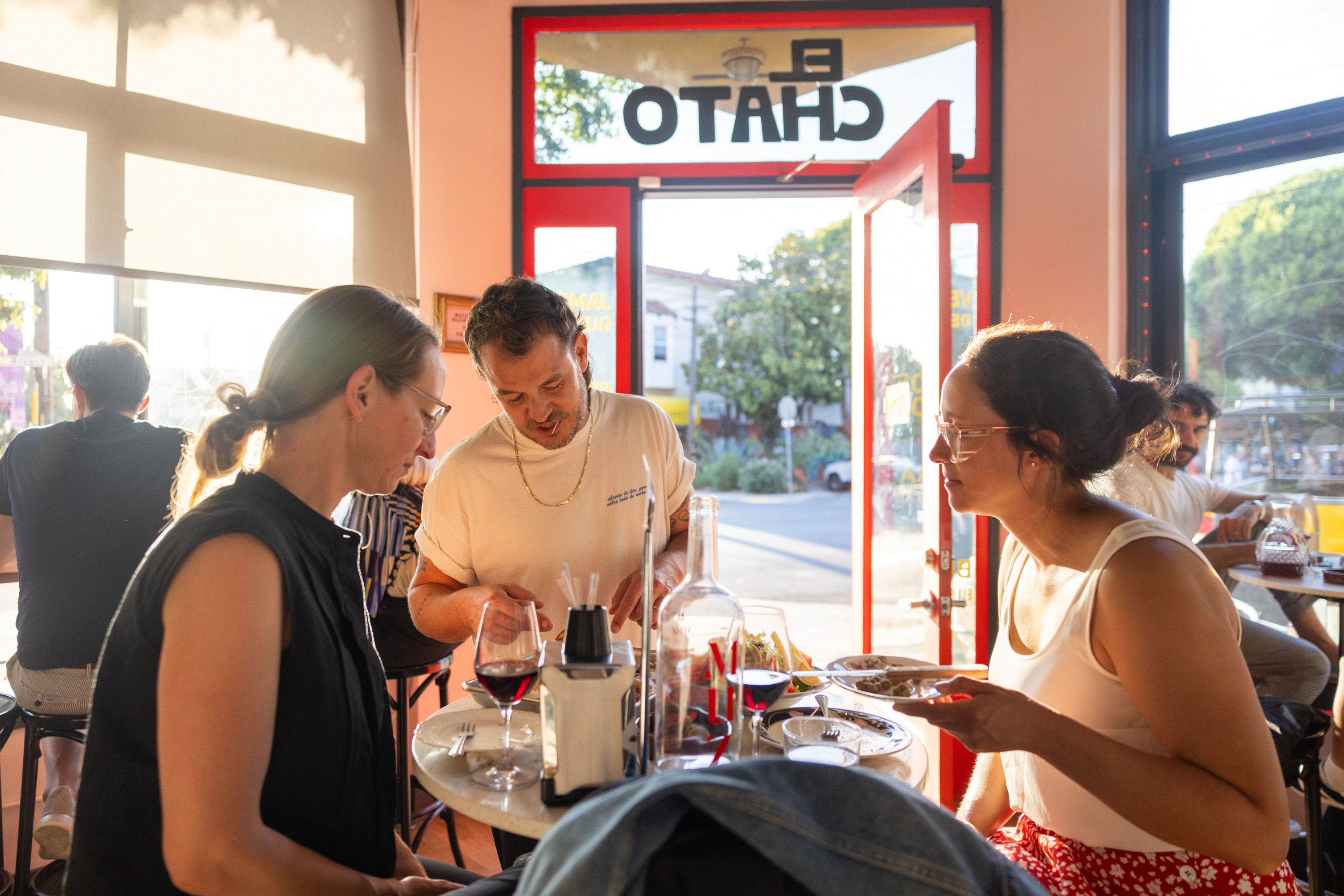 Three people are chatting around a table in a café, with sunlight streaming in through open doors. There are drinks and plates of food in front of them.