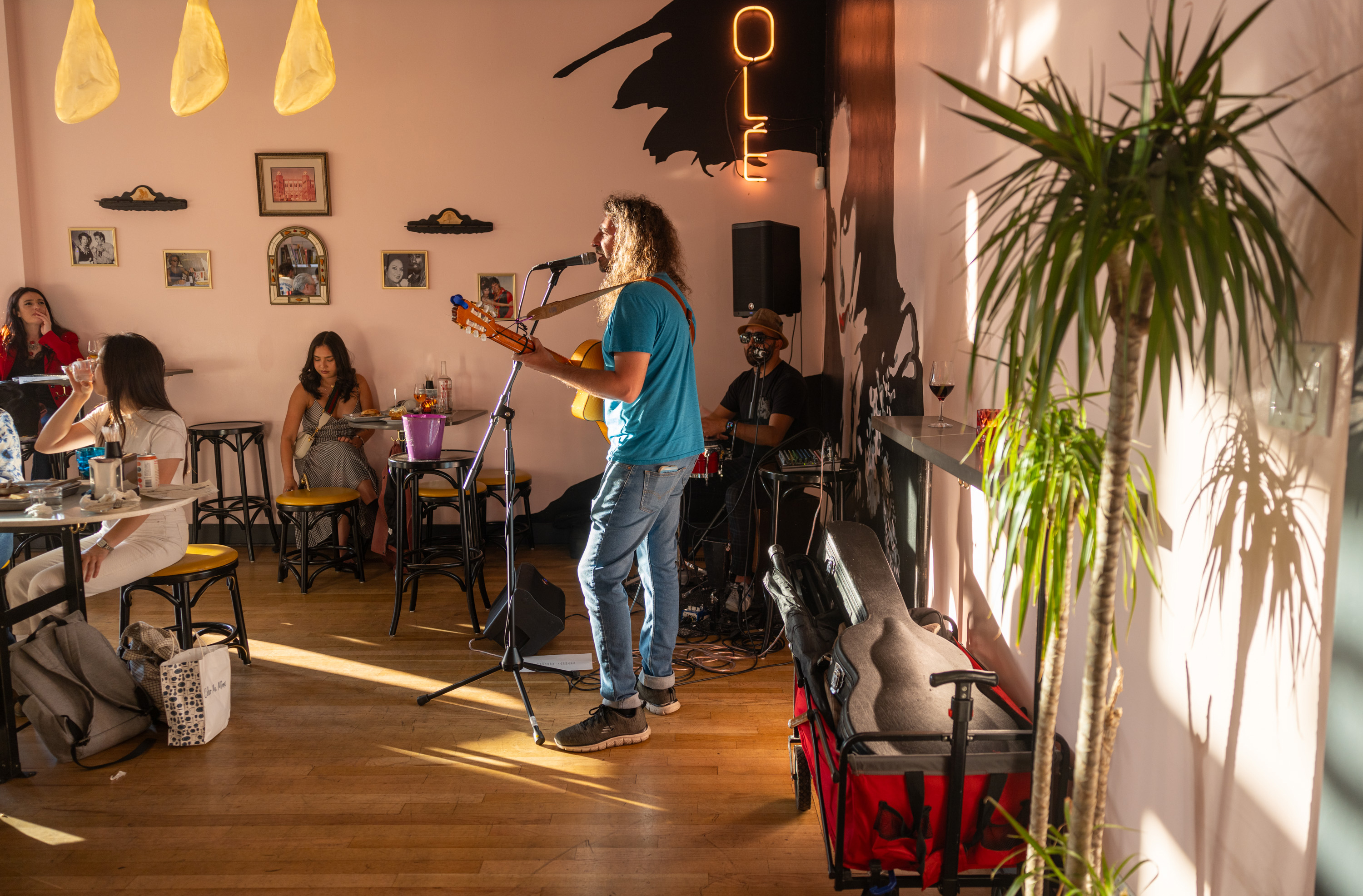 A musician in a blue shirt plays the guitar in a cozy cafe, while people seated at tables watch. Framed pictures decorate the pink walls, and a neon sign says &quot;OLE.&quot;