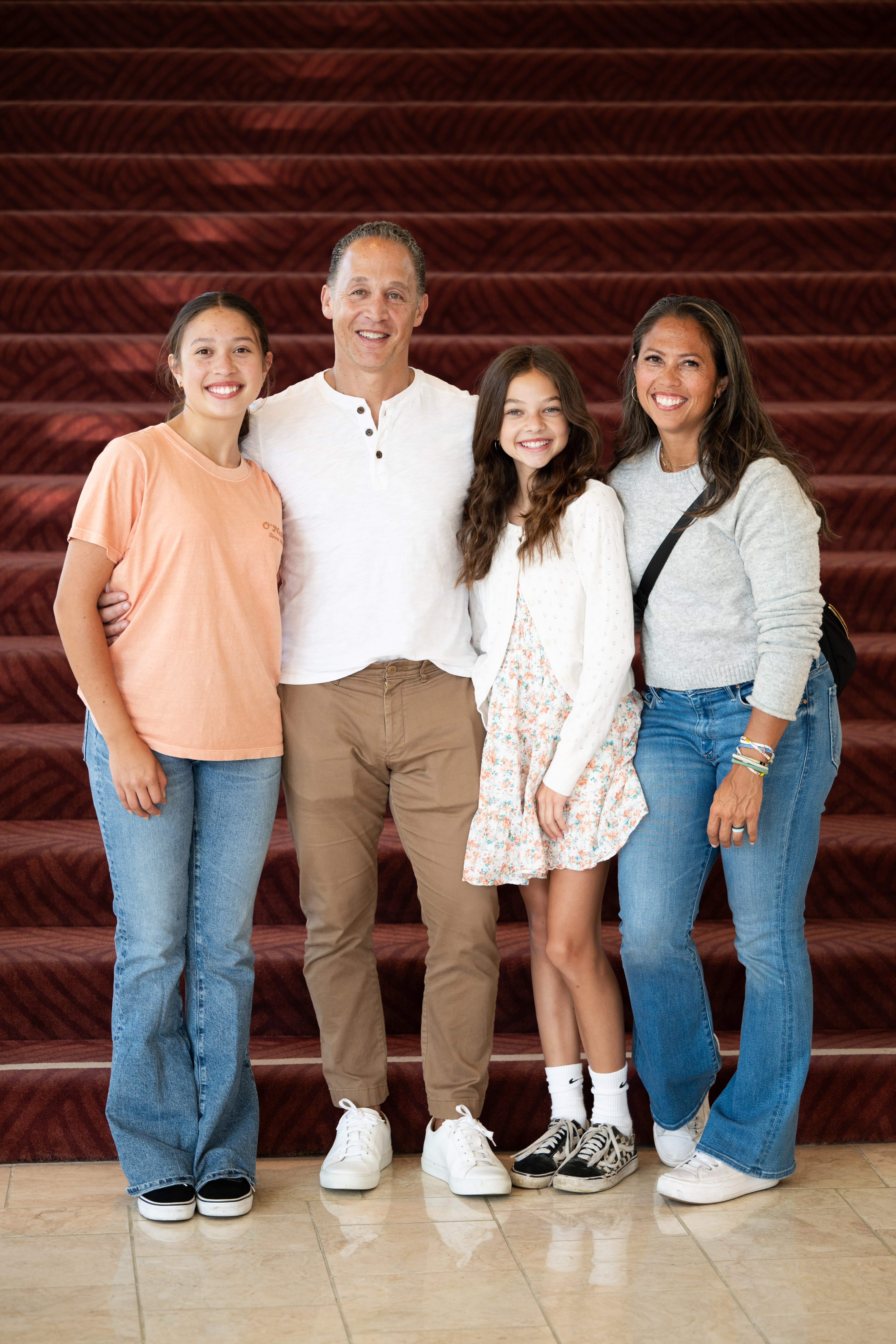 A family of four stands smiling in front of red-carpeted stairs. The two girls wear casual outfits while the parents are dressed in semi-casual attire.