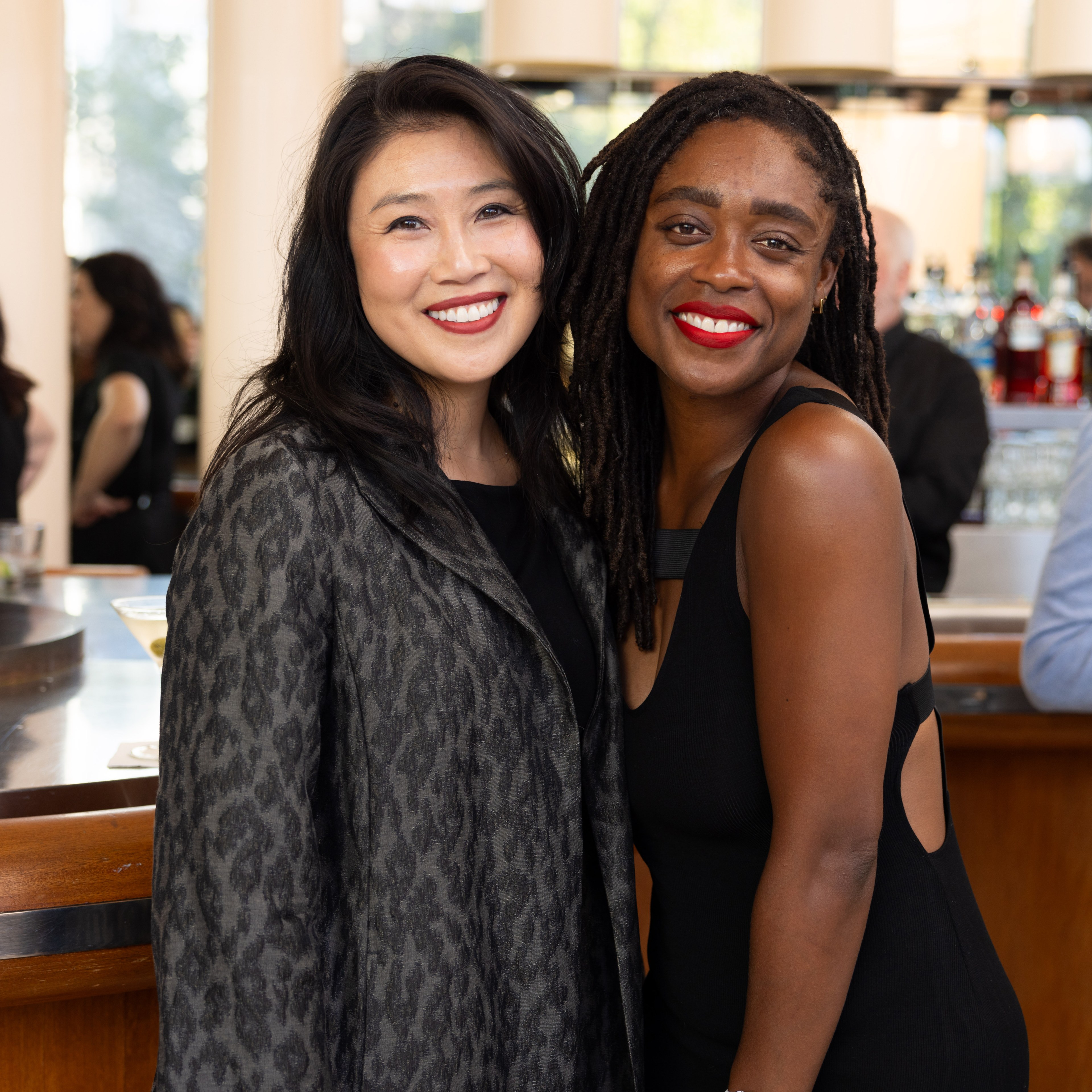 Two women are smiling and posing together; one has long dark hair and wears a gray patterned jacket, while the other has long braided hair and wears a black dress.