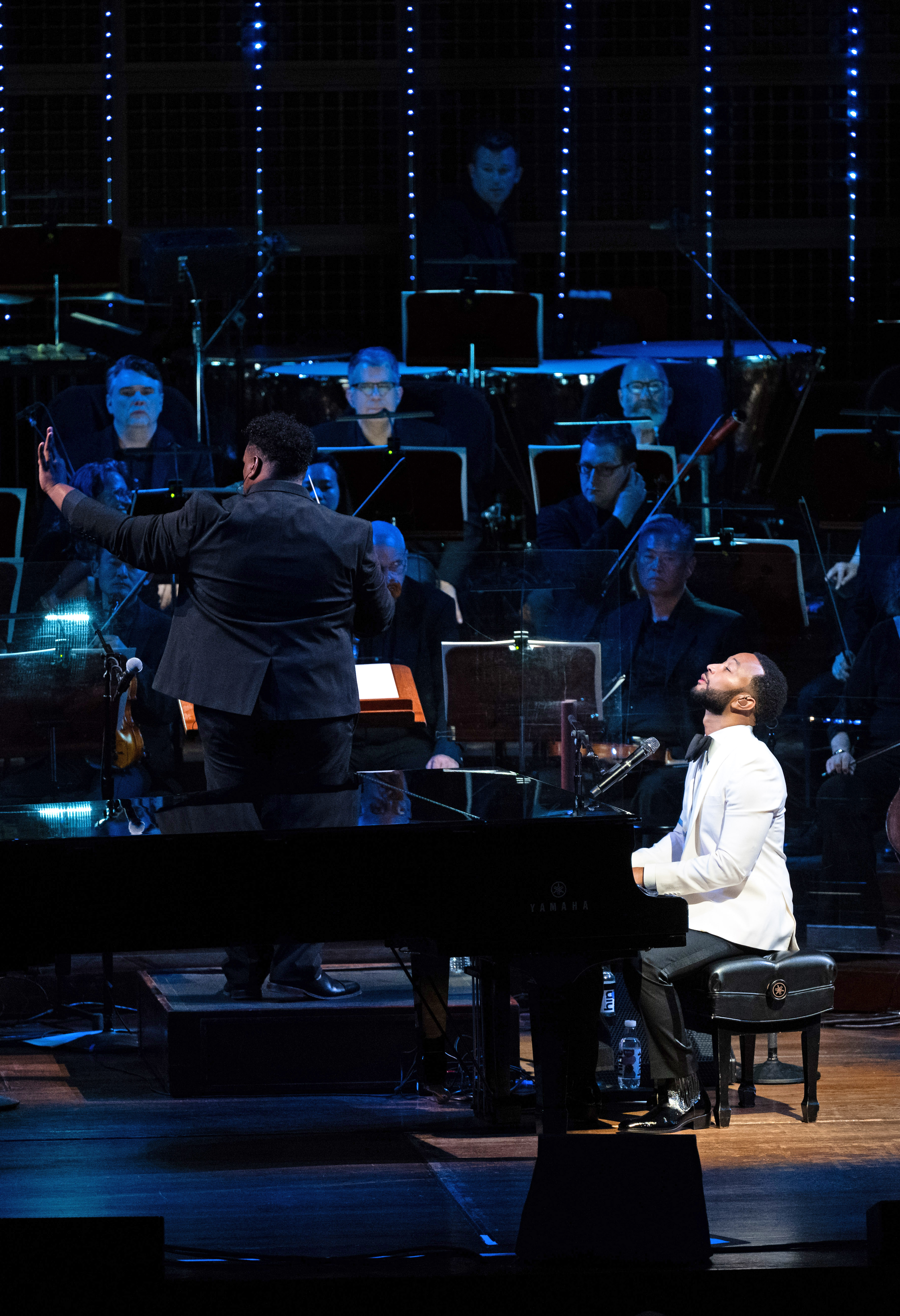 A pianist in a white jacket plays while looking upwards on stage beside a grand piano. A conductor in a black suit directs an orchestra in the background.