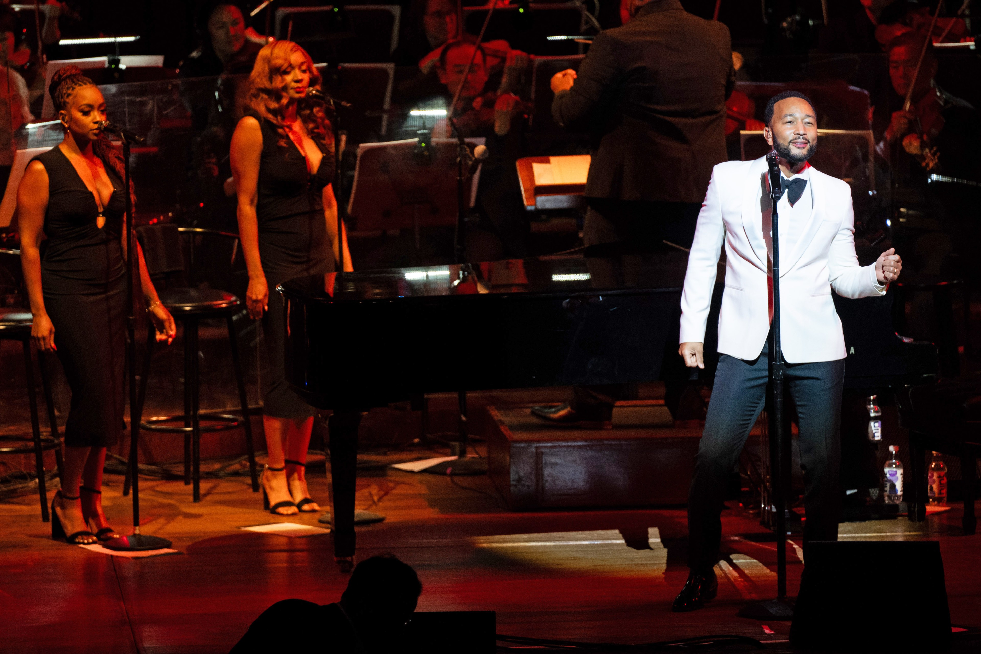 A man in a white jacket performs on stage, singing into a microphone, with two women in black dresses as backup singers, accompanied by an orchestra in the background.