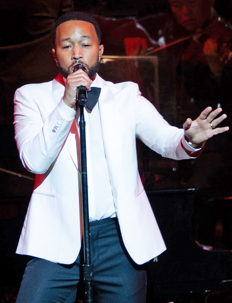 A man in a white suit jacket with a black bow tie sings passionately into a microphone. He has a beard and is standing on stage, illuminated under a spotlight.