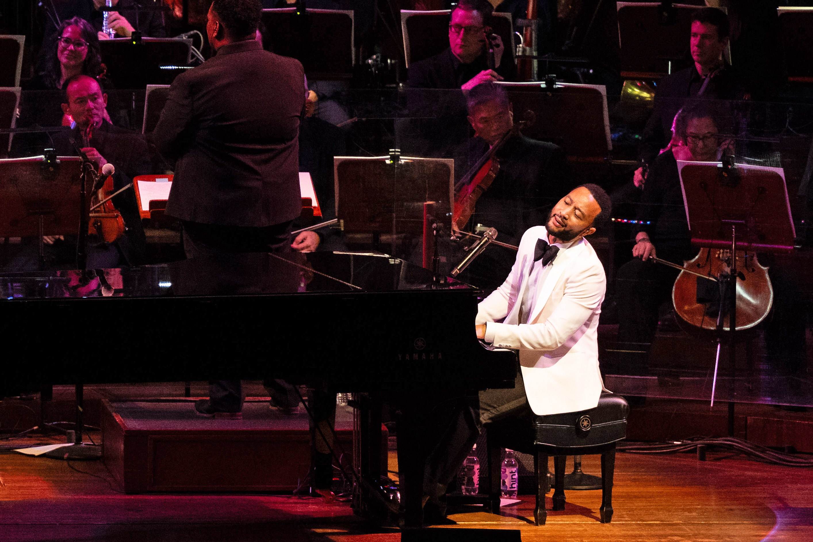 A man in a white tuxedo jacket plays a grand piano on stage, eyes closed. Behind him, an orchestra of musicians in black attire performs under the direction of a conductor.