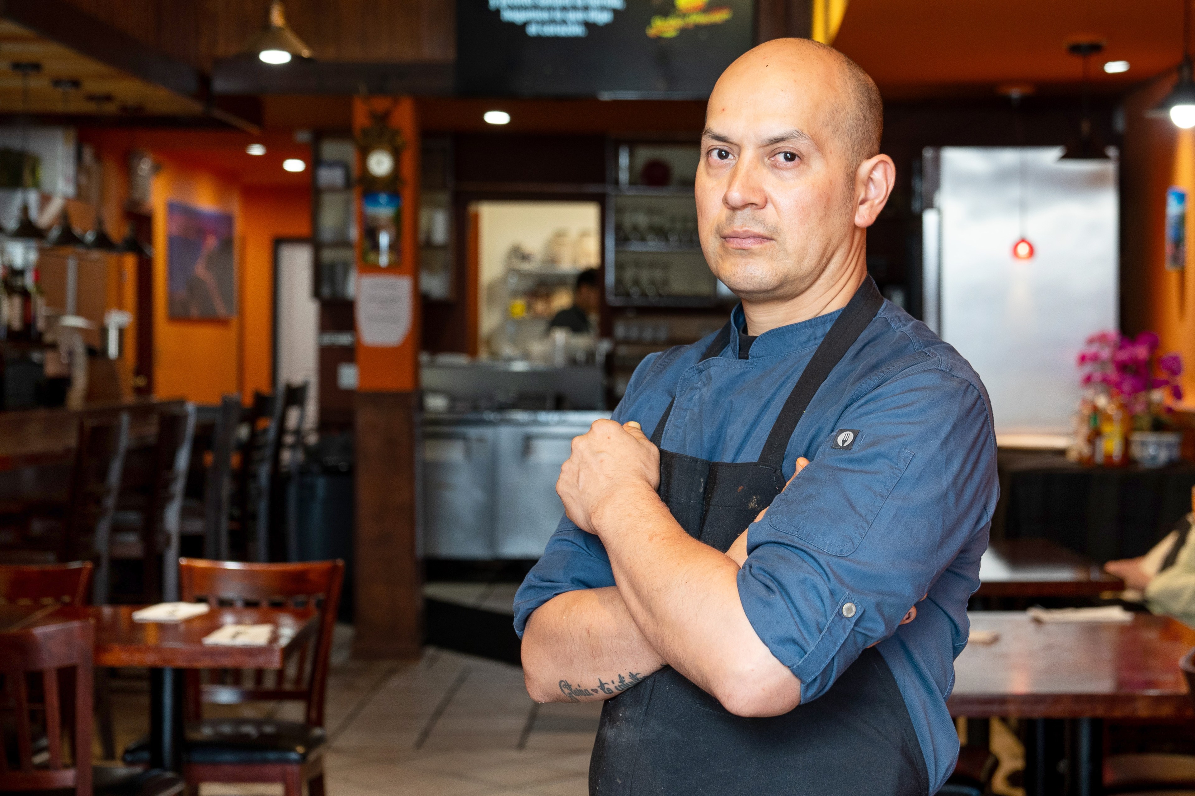 A man wearing a blue chef coat and black apron stands confidently in a warmly lit restaurant, which features wooden chairs, tables, and orange walls.