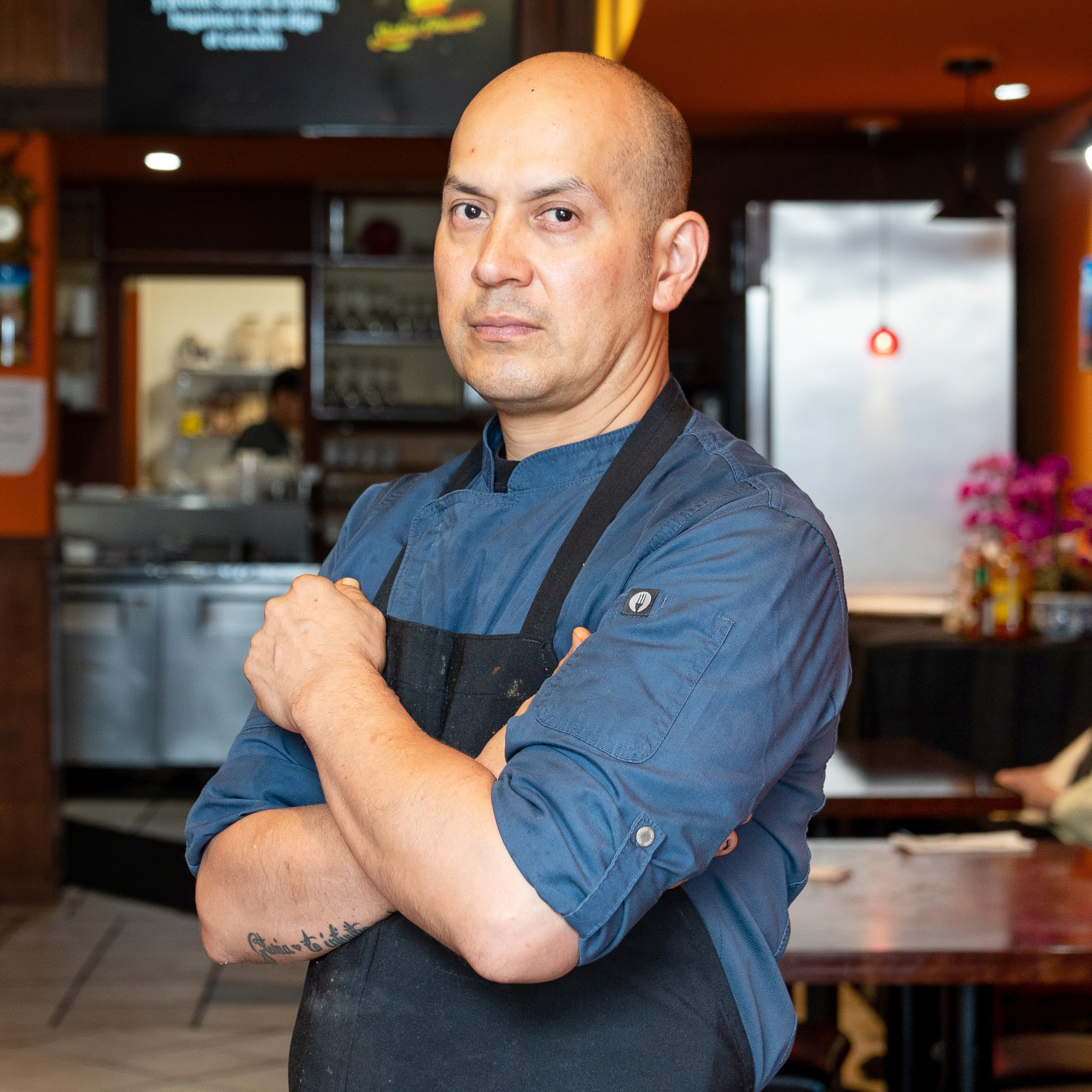 A man wearing a blue chef coat and black apron stands confidently in a warmly lit restaurant, which features wooden chairs, tables, and orange walls.