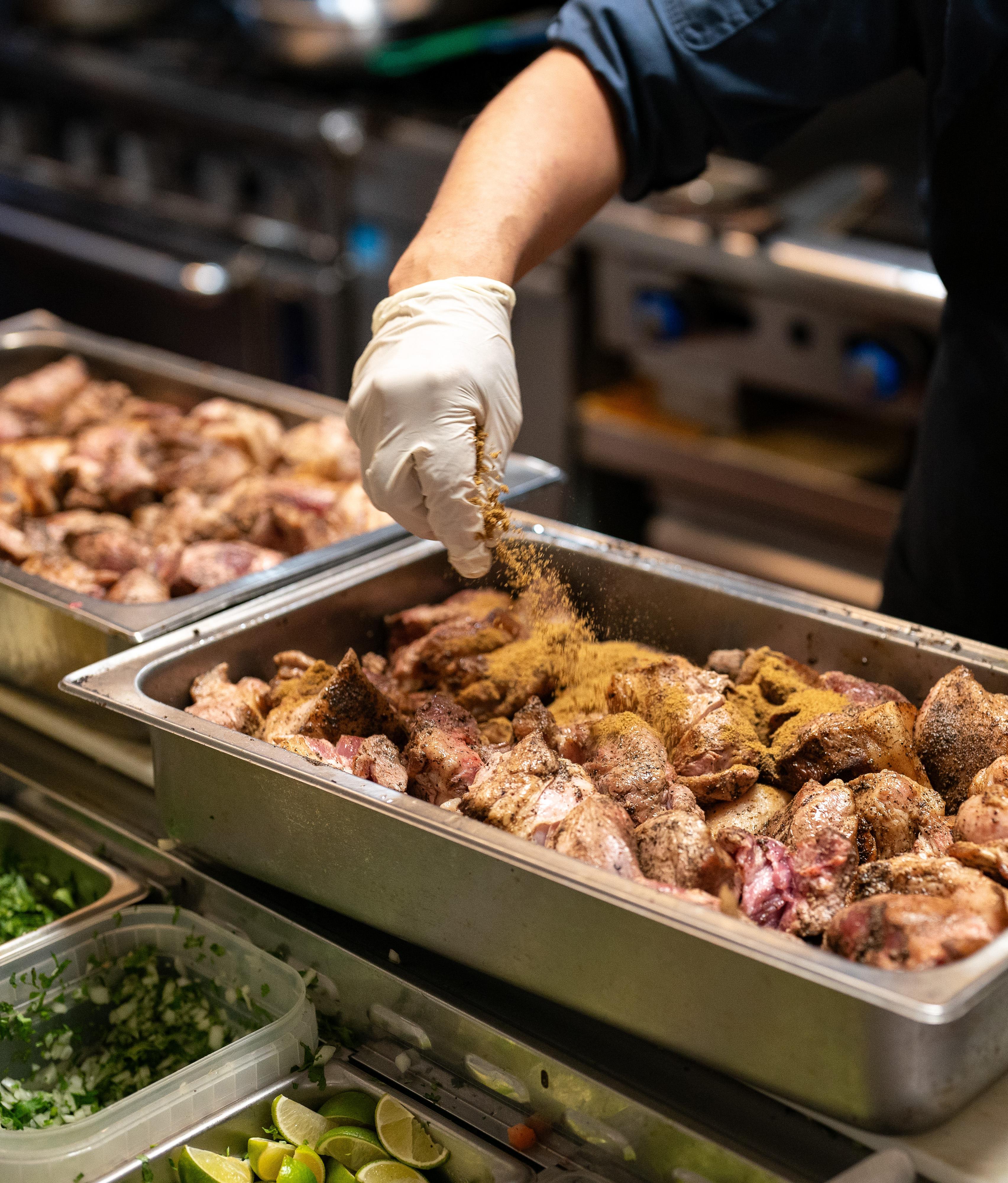 A person wearing a glove is sprinkling seasoning over raw meat in a metal tray in a kitchen, with chopped herbs and lime wedges visible in nearby containers.